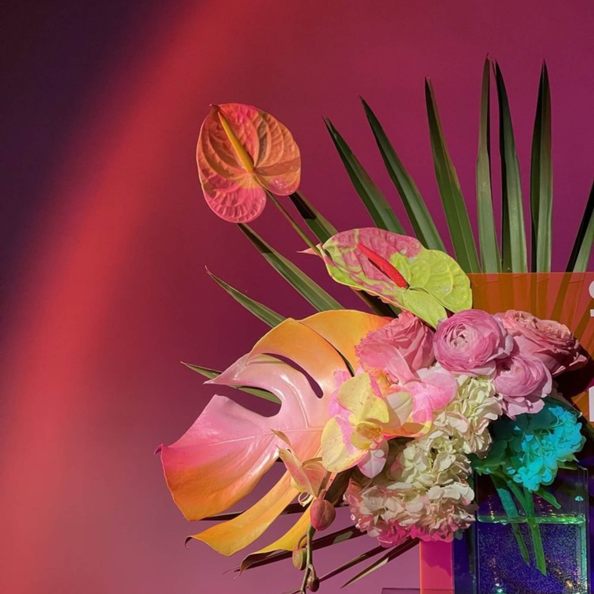 Tropical floral arrangement with pink anthuriums, orchids, and palm leaves in a clear vase