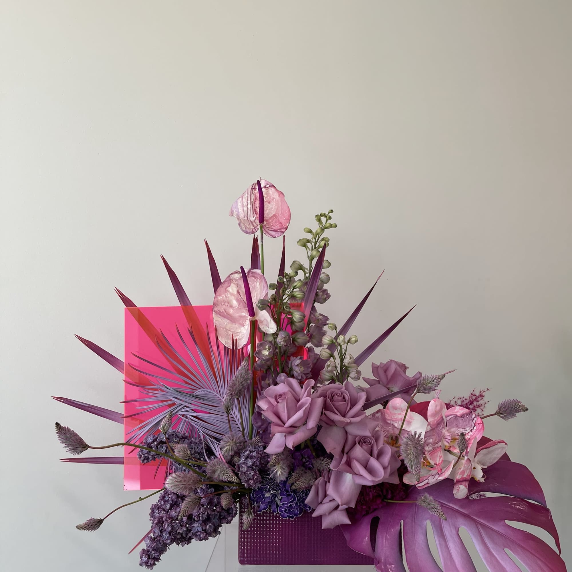 Purple floral arrangement with roses, orchids, and tropical leaves in a pink container