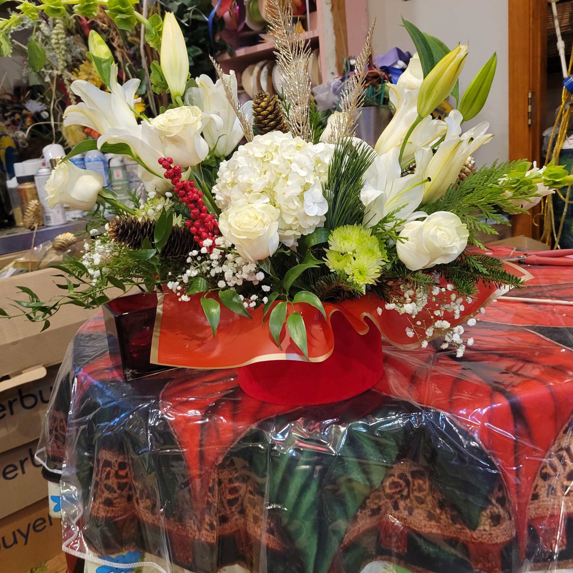 White lilies and roses arranged in a red hatbox with holiday accents.