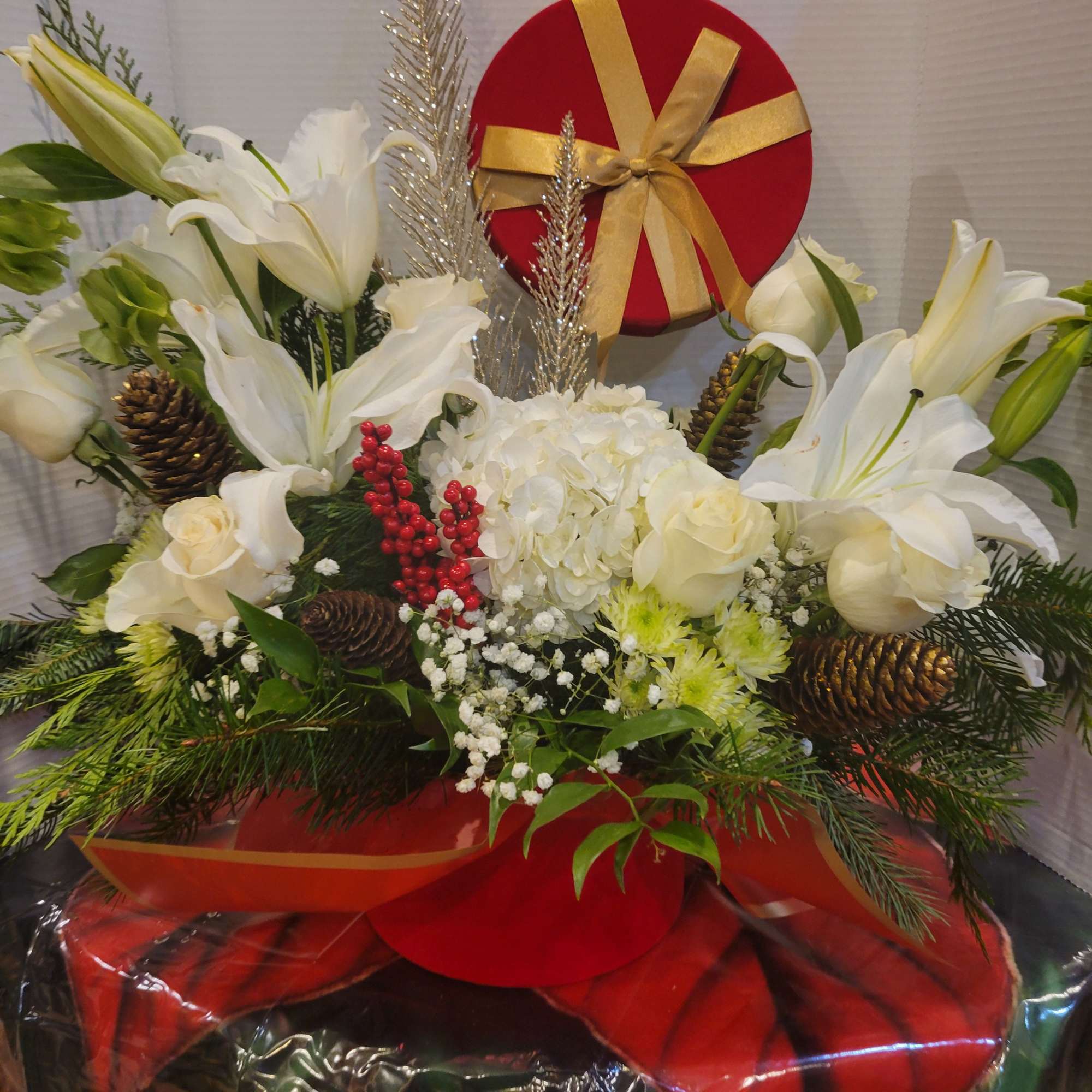 White lilies and roses arranged with a red gift box and gold ribbon