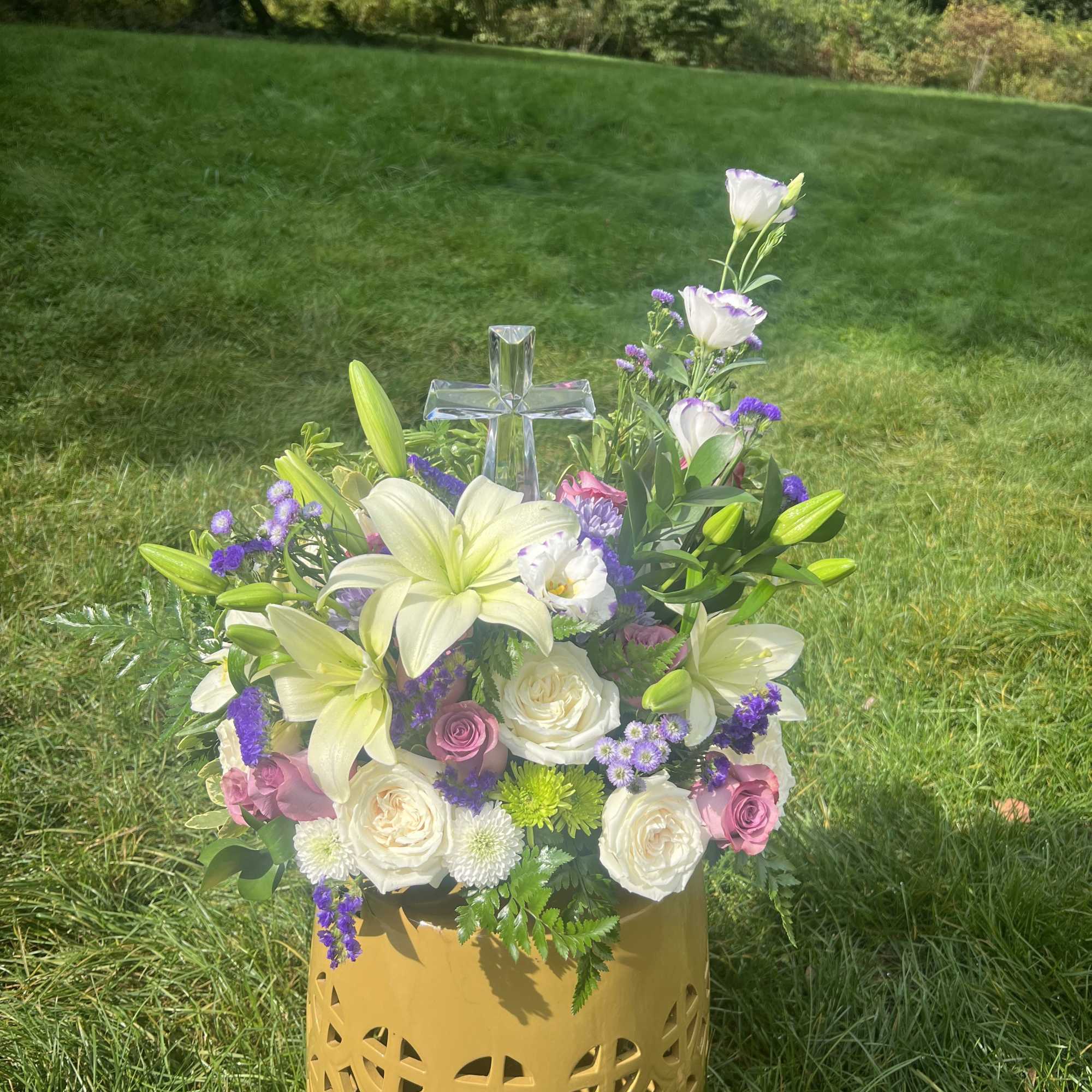 Floral arrangement with white lilies, roses, and purple accents in a gold vase with a clear cross