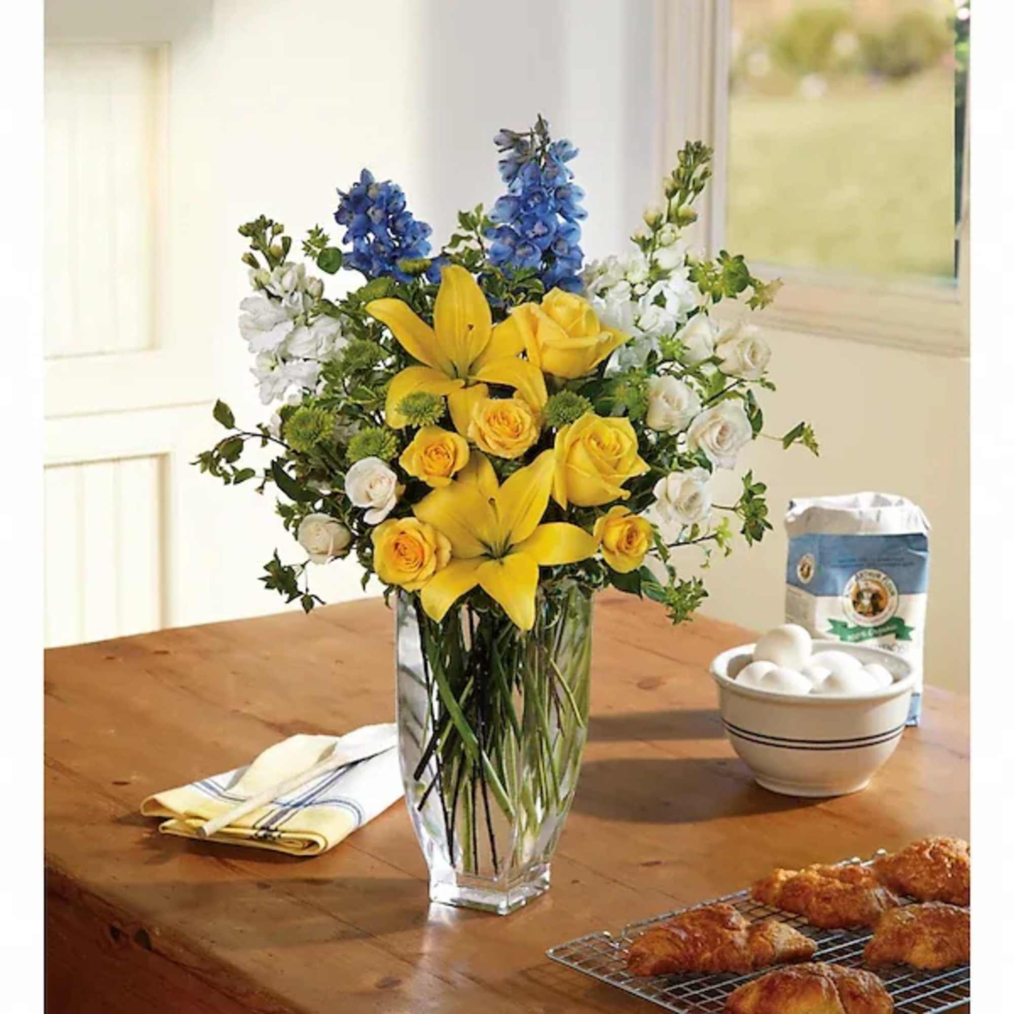 Yellow lilies, roses, and blue flowers in a clear glass vase