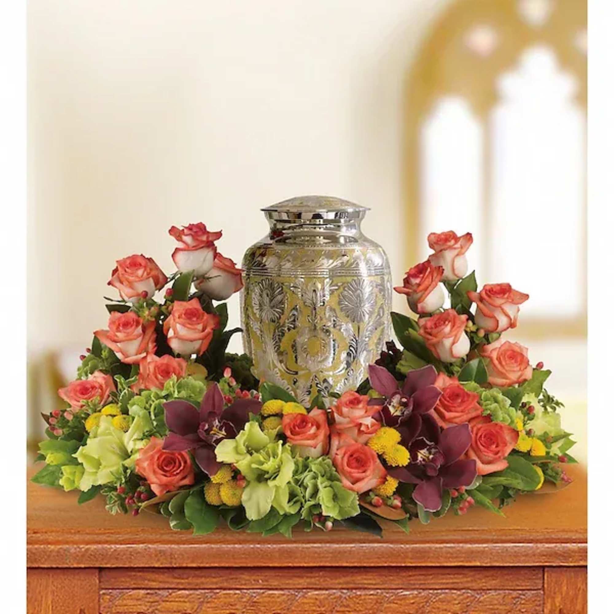 Floral urn arrangement with coral roses and green blooms around a silver vase
