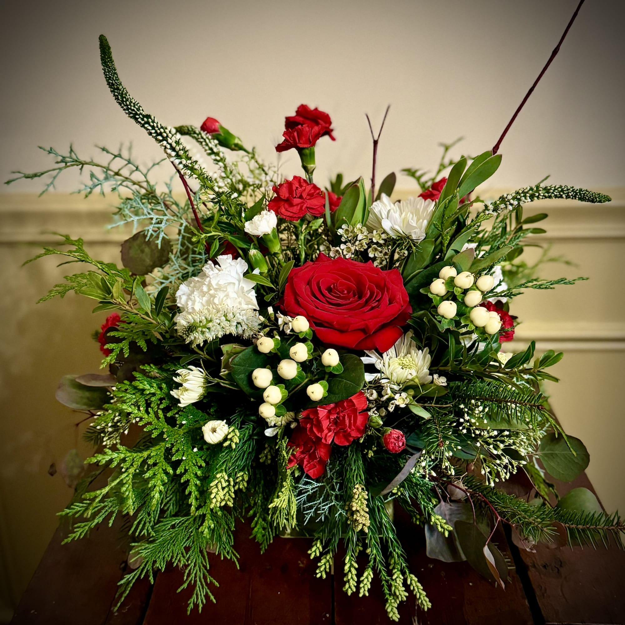 Low arrangement of red roses, red and white flowers, and berries with abundant evergreen branches.