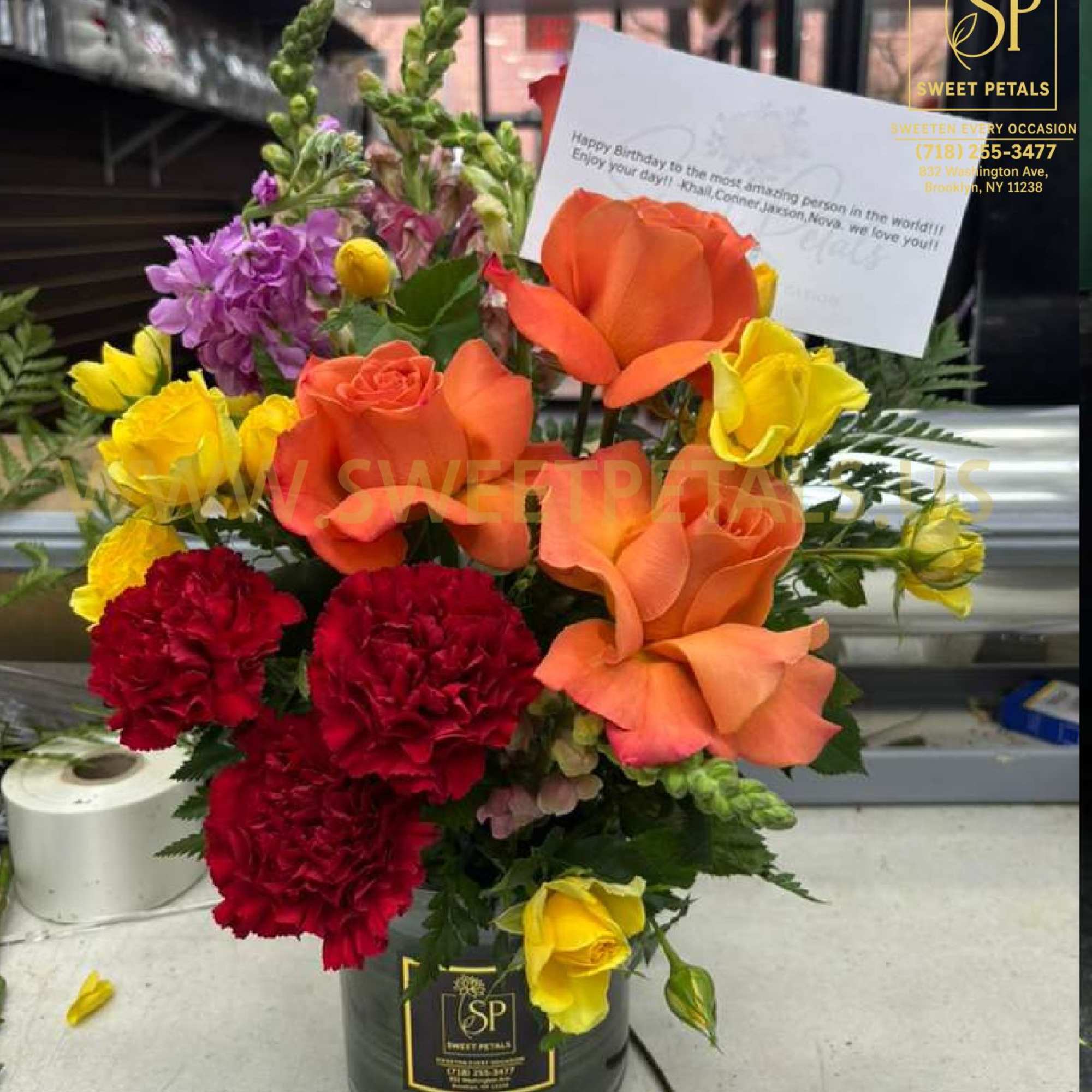 Mixed bouquet with orange roses, yellow roses, red carnations, and purple flowers in a black container