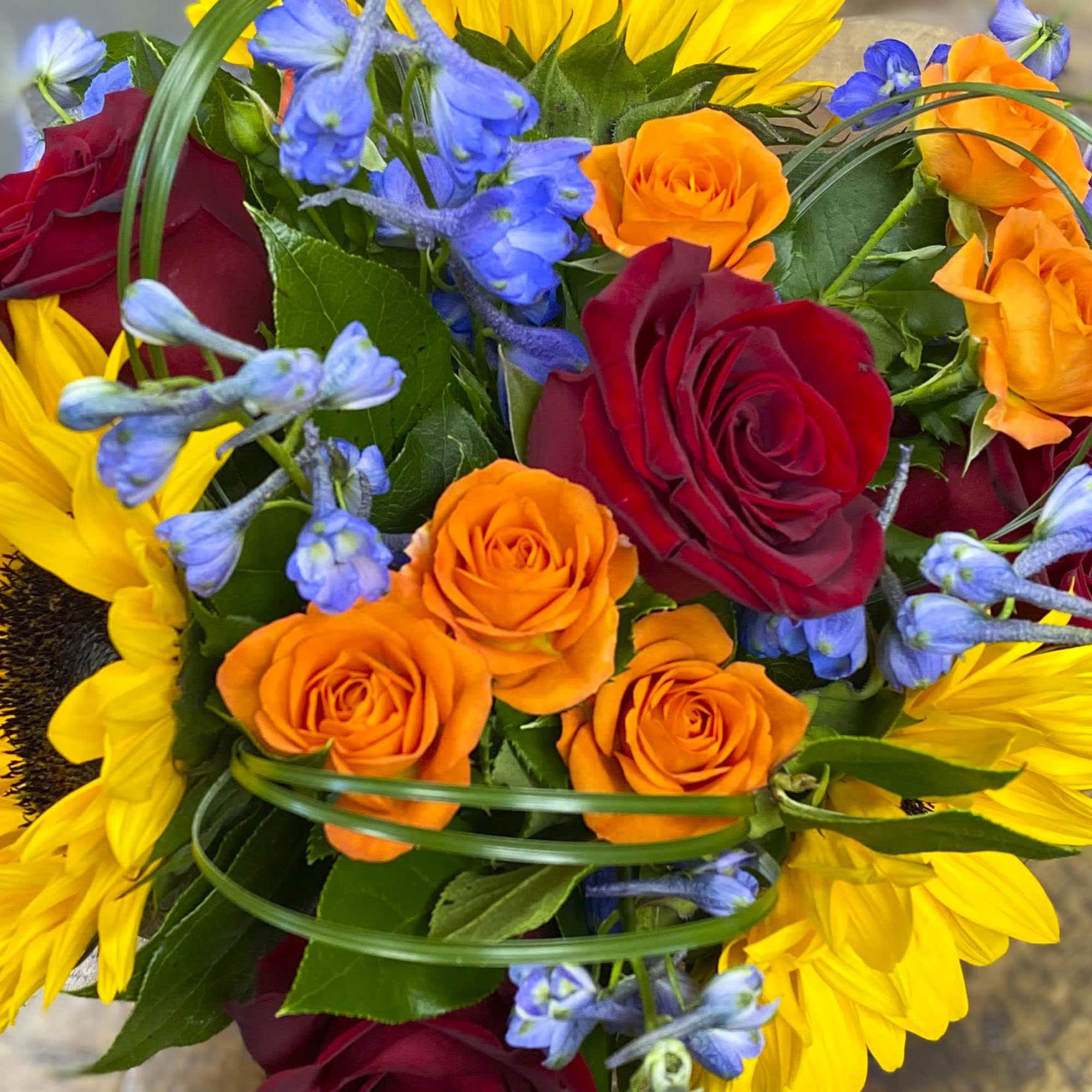 This beautiful arrangement is filled with sunflowers, roses, garden roses, blue delphiniums