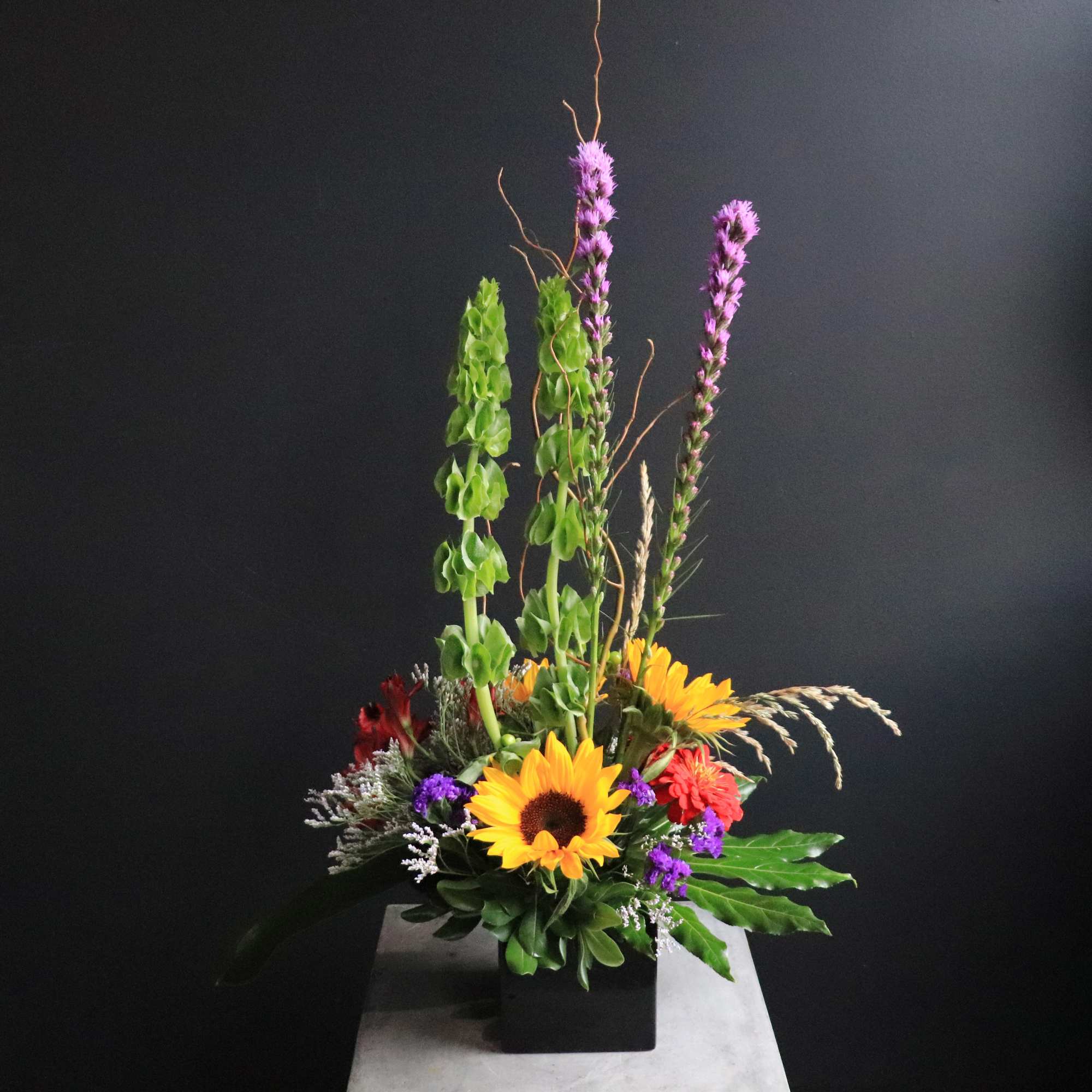 In a modern black container:  Statice, Sunflowers, Aspidistra, Mums, Curly Willow