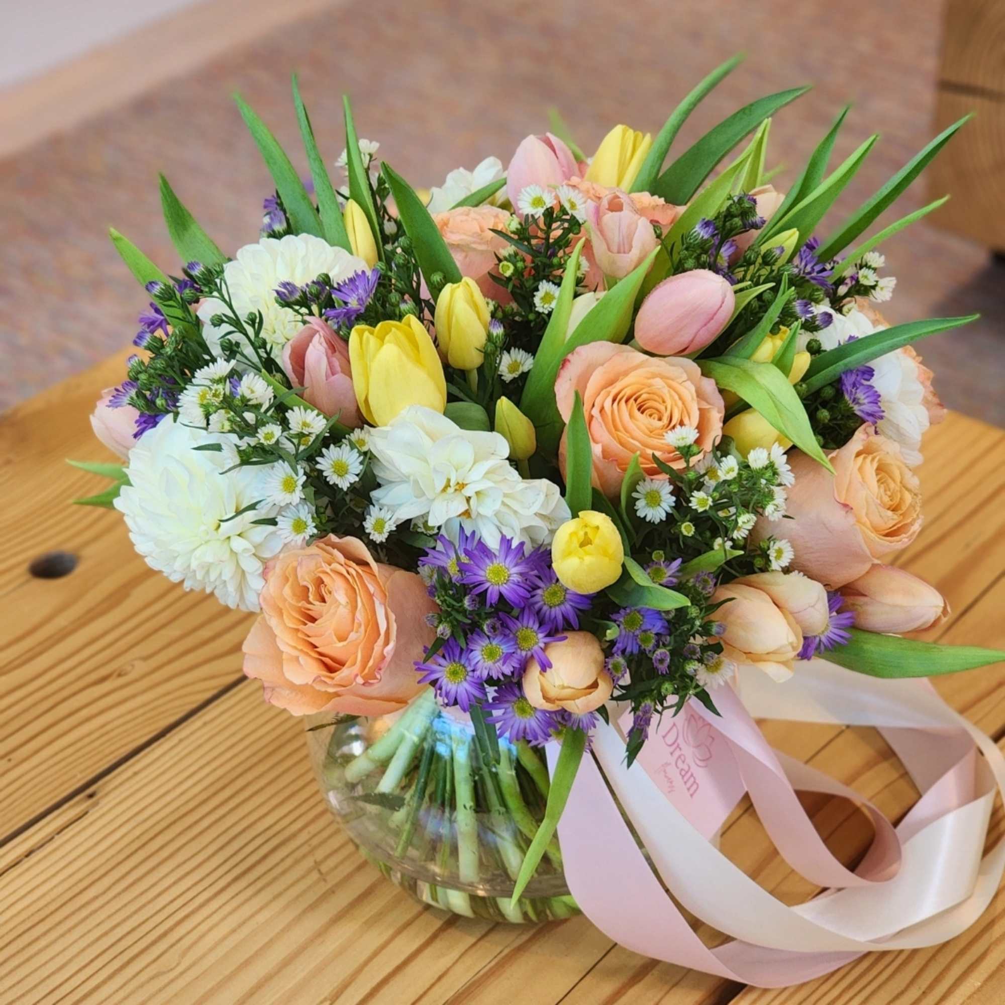  A clear vase arrangement featuring peach roses, yellow tulips, white dahlias