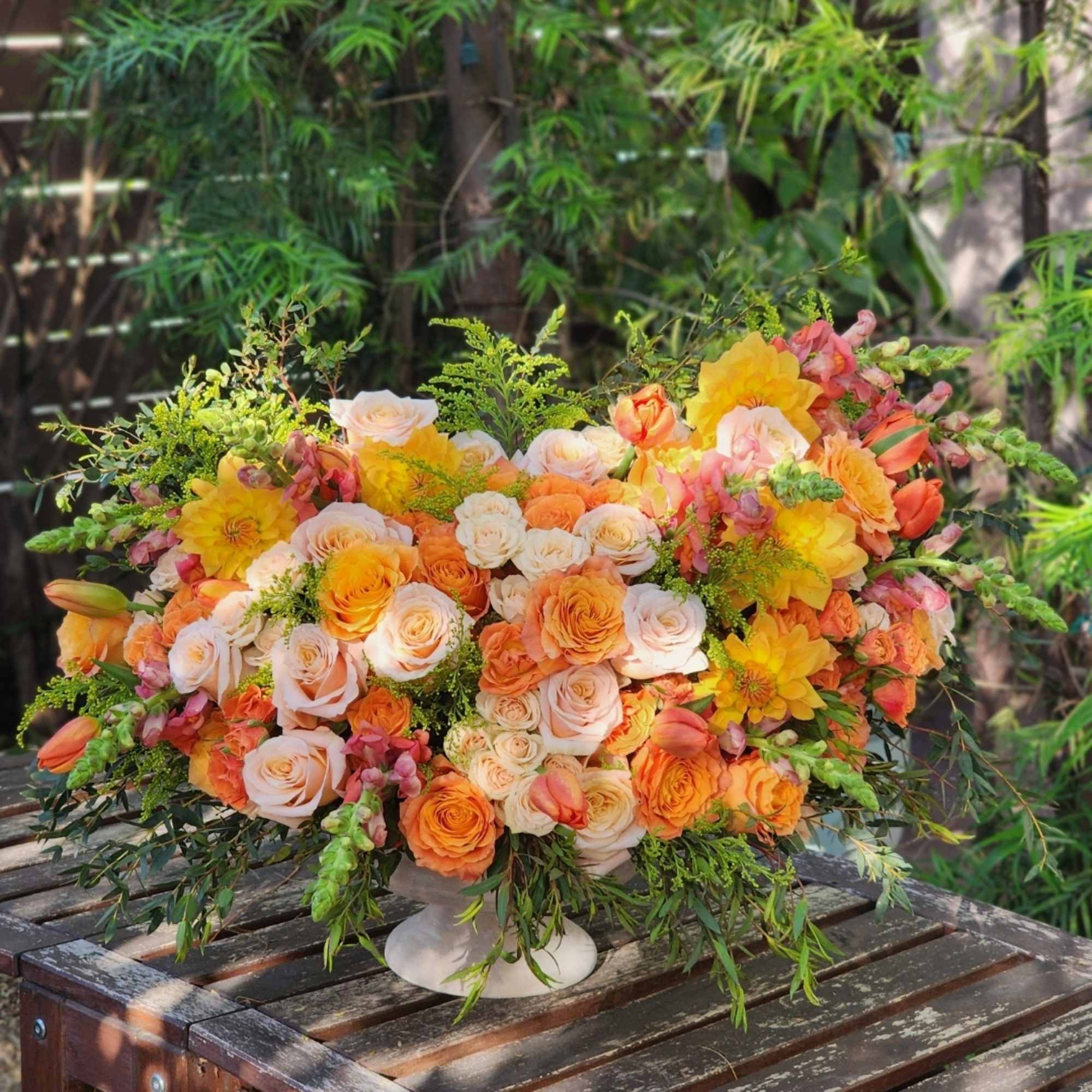 The arrangement features vibrant oranges dahlias, soft peaches roses, and lush greens