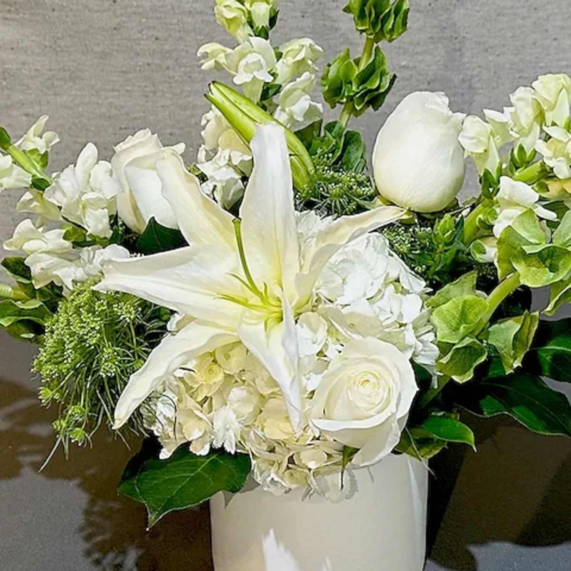 This calming arrangement of all white and green seasonal flowers, roses, lilies