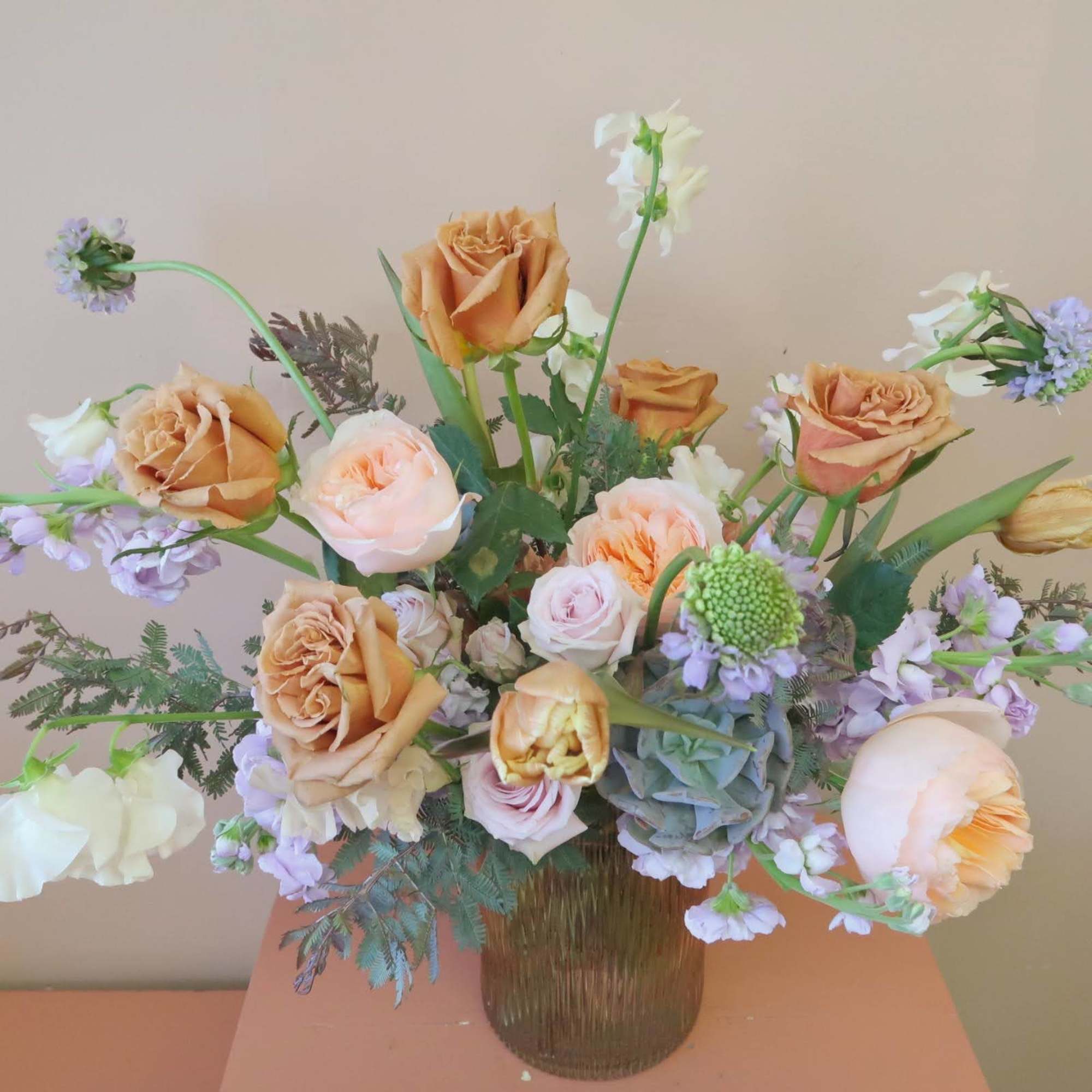A soft hued arrangement featuring trendy toffee roses and lavender hues with