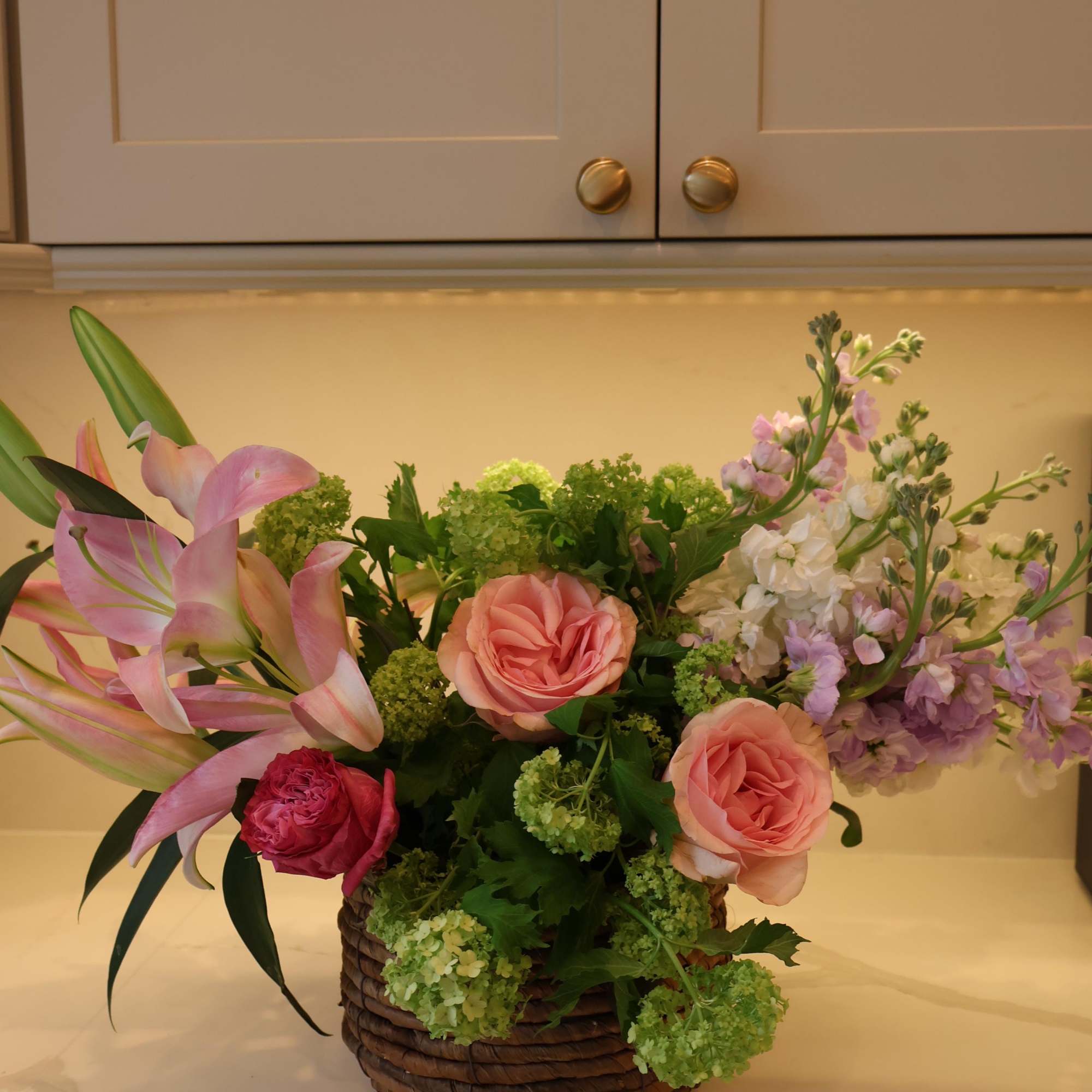 A charming basket of blush roses, pink lilies, lavender stock, and fresh