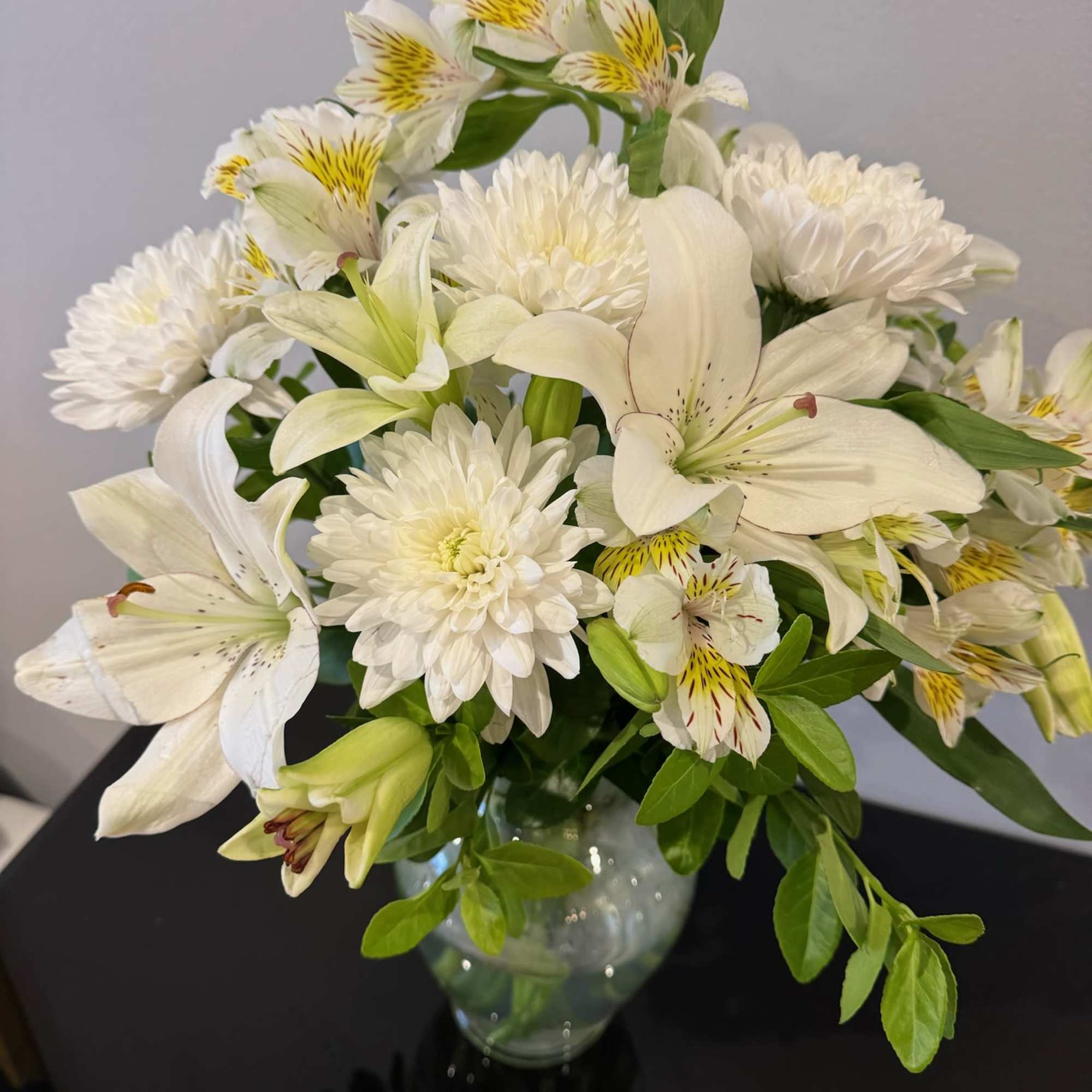 A sympathy arrangement featuring White Mums, White Lilies, Alstroemeria, and assorted greens