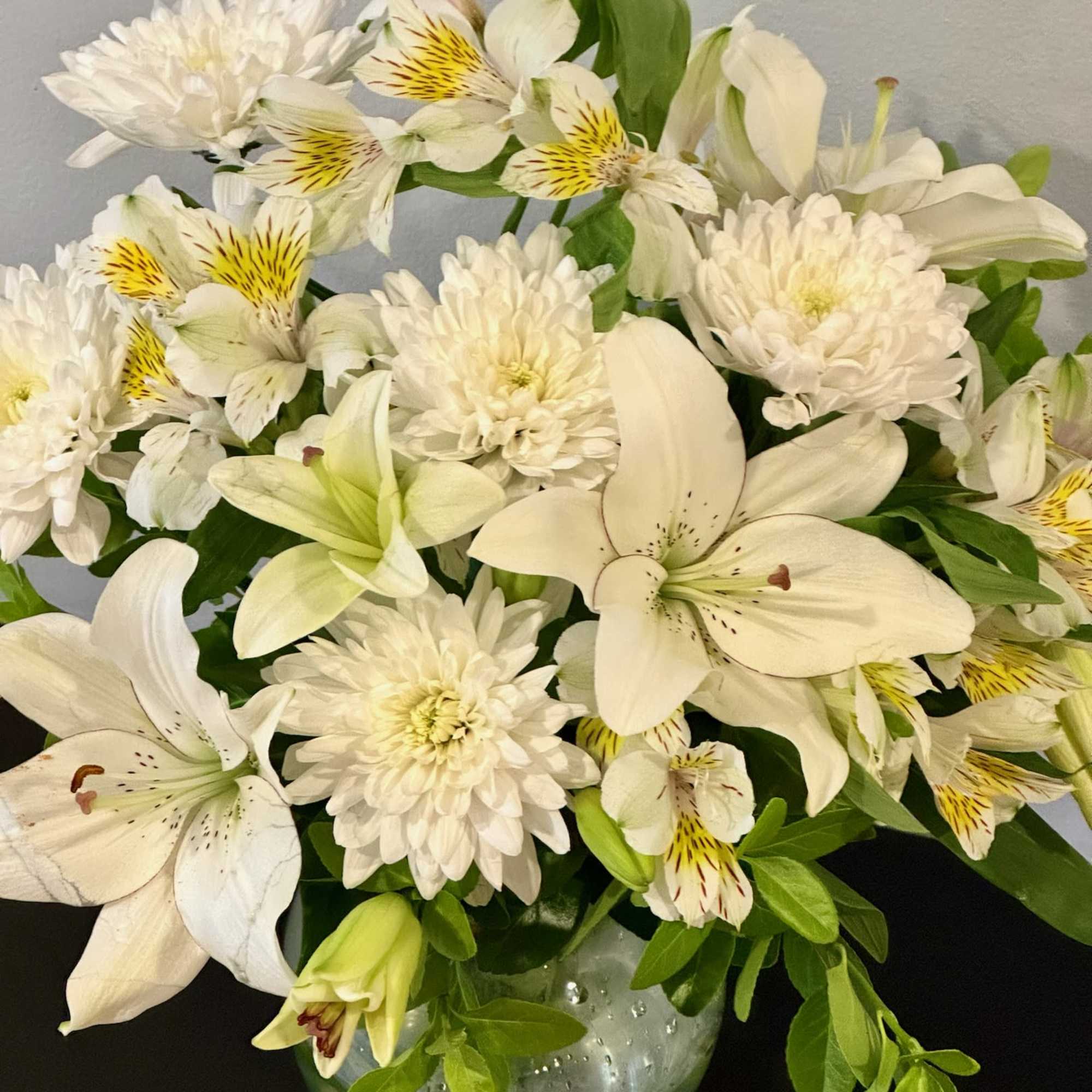 A sympathy arrangement featuring White Mums, White Lilies, Alstroemeria, and assorted greens
