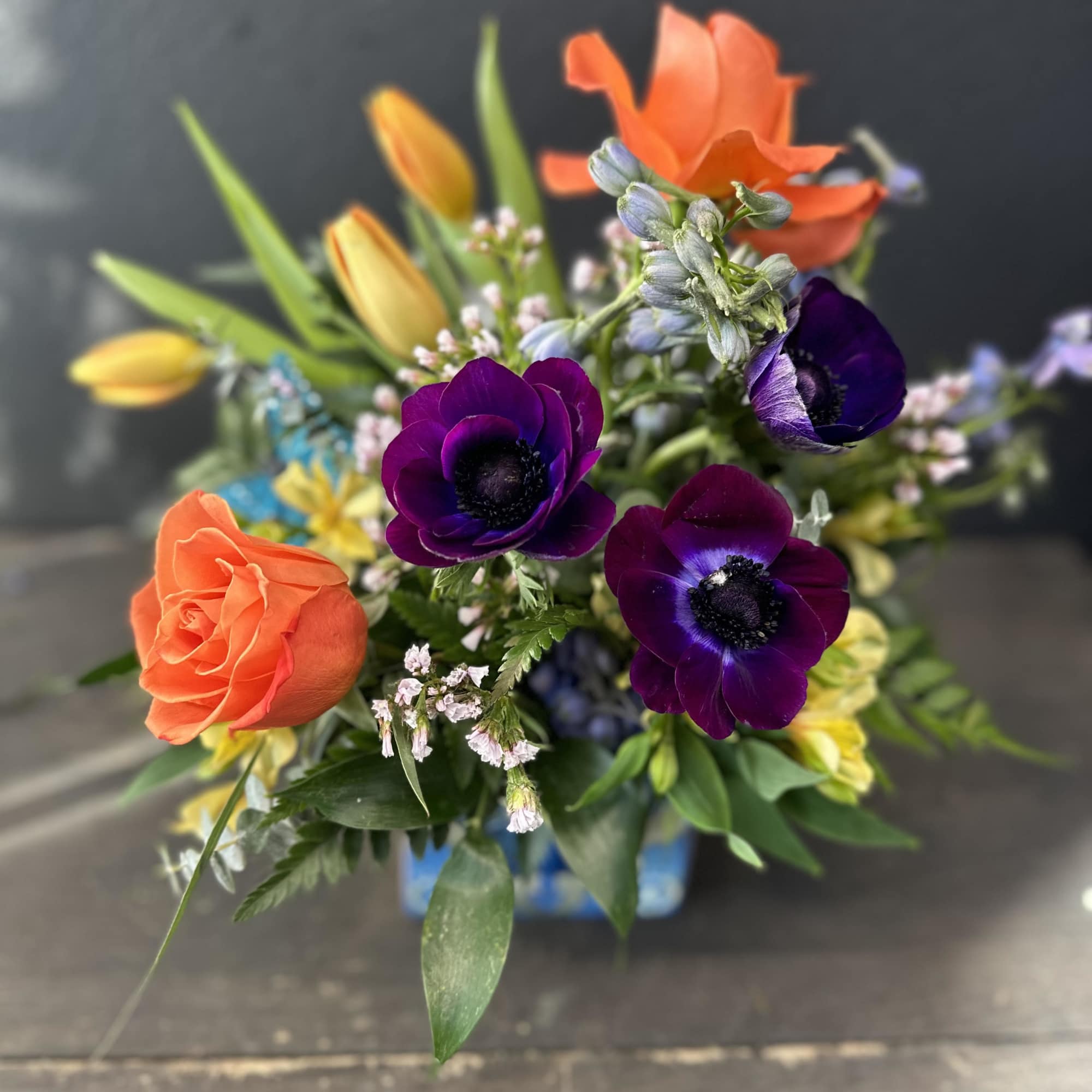 Designed using plum anemones, golden yellow tulips, reflexed orange roses, light blue