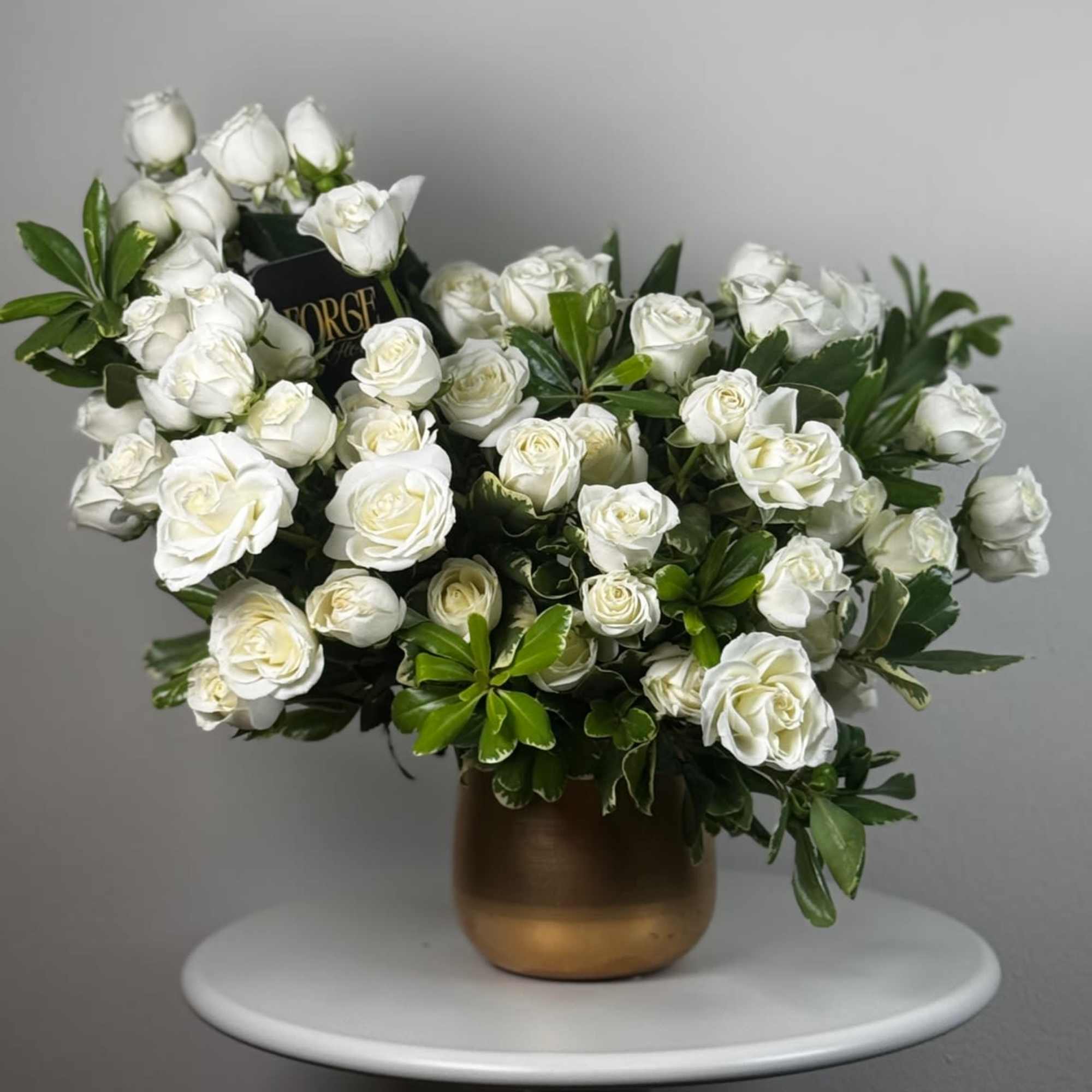 Bold white spray roses arranged with lush greenery in a matte-gold vase.