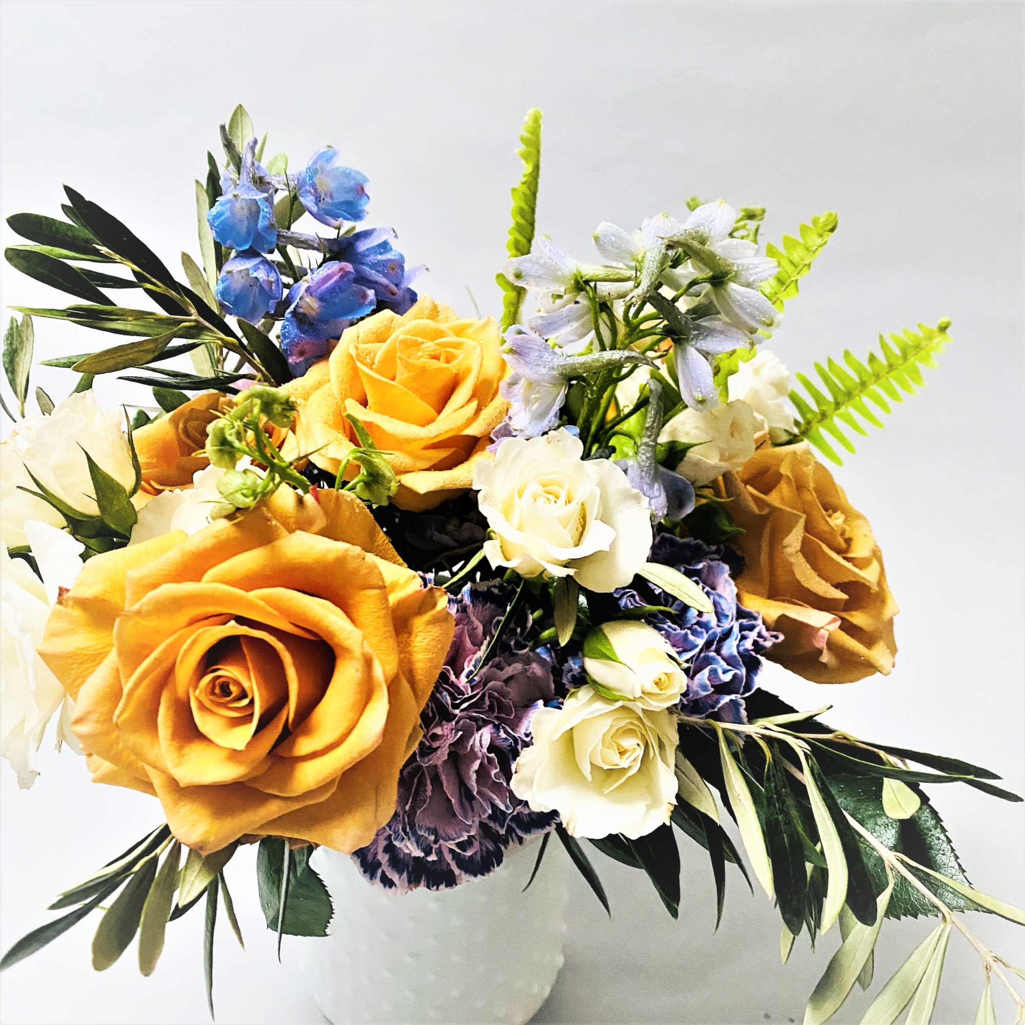 Comprised of gold mustard roses, white spray roses, blue delphinium with mixed