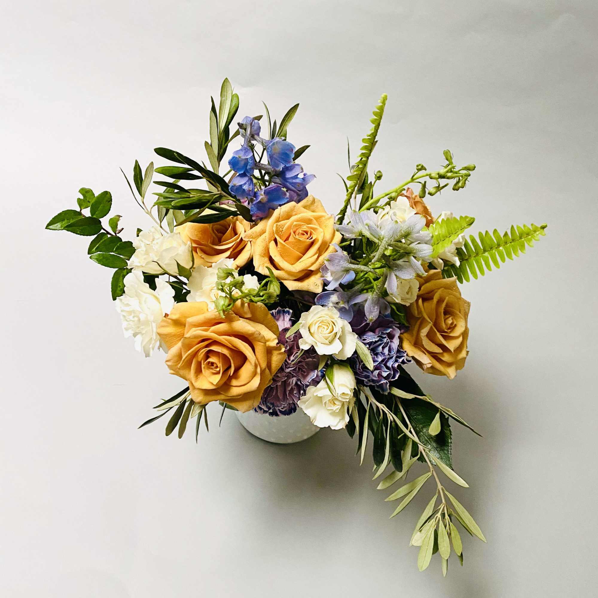 Comprised of gold mustard roses, white spray roses, blue delphinium with mixed
