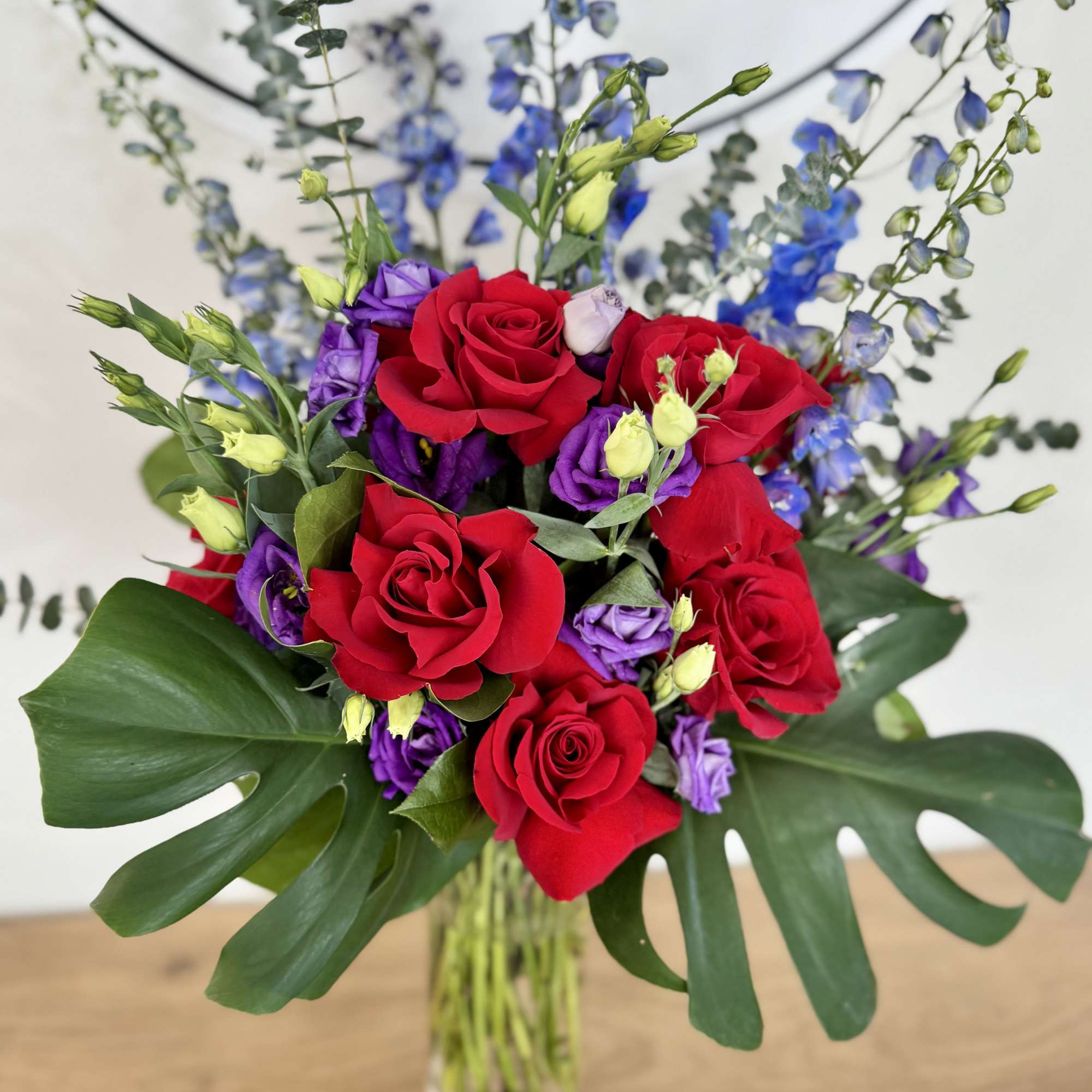 Featuring monstera leaves, delphinium, roses, and eucalyptus, this blue, green, and red