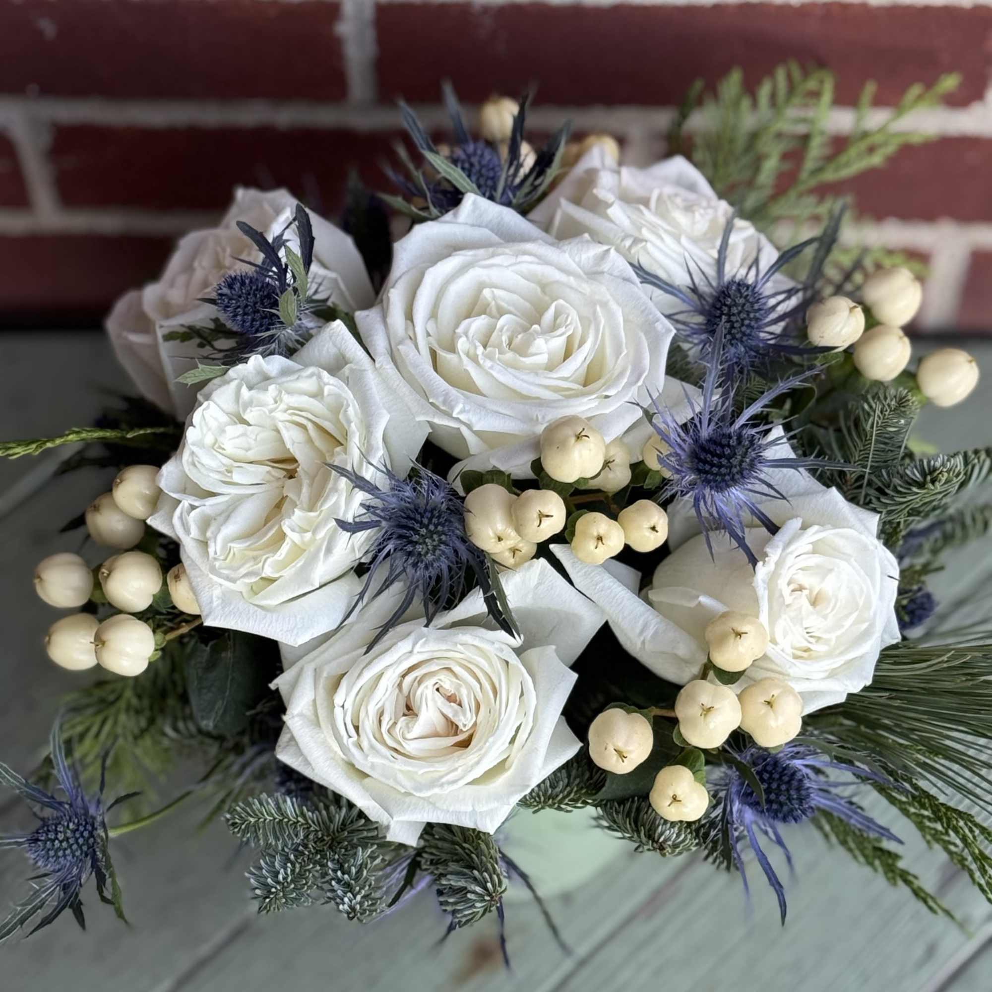 We arrange white roses with hypericum and thistle with mixed evergreens. Pot