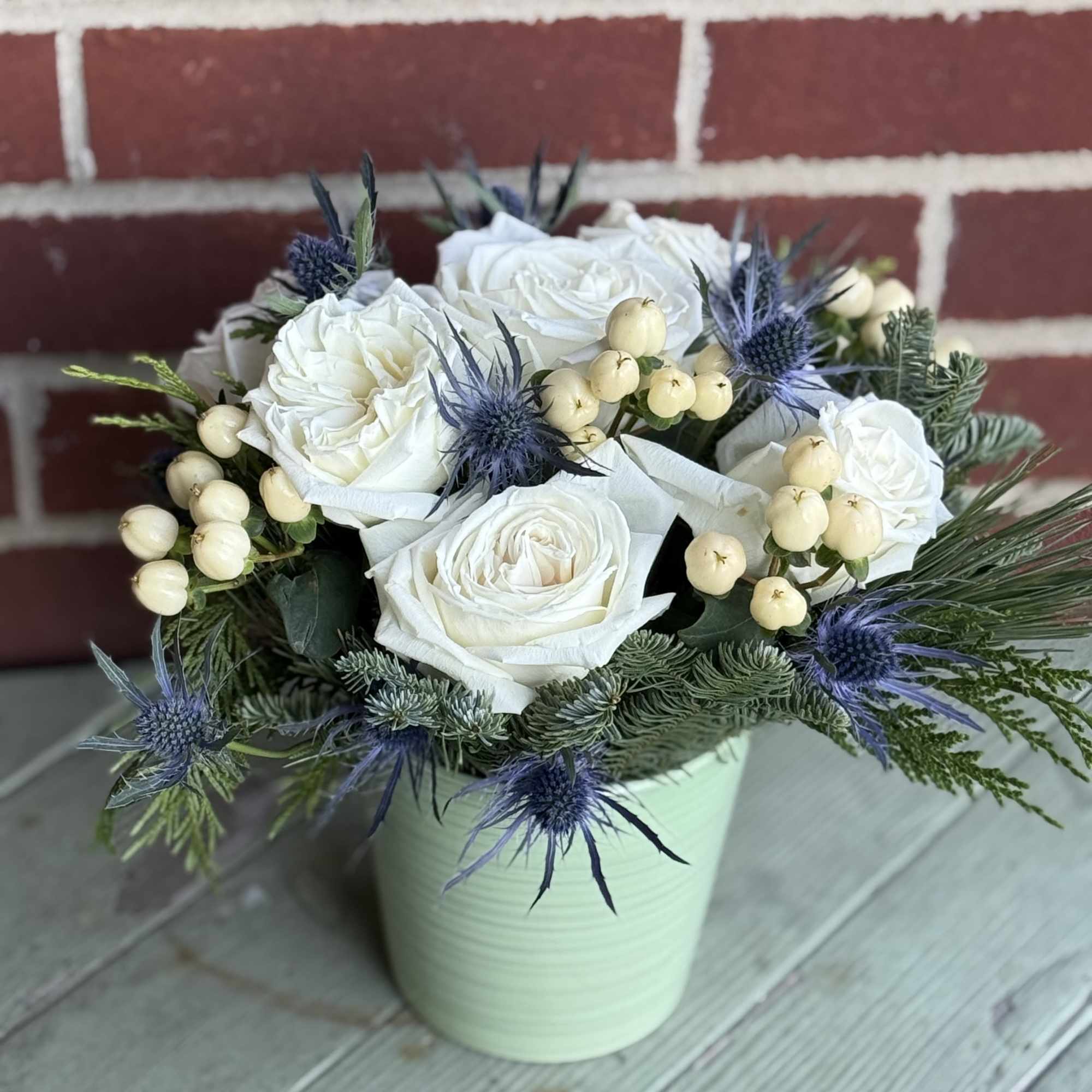 We arrange white roses with hypericum and thistle with mixed evergreens. Pot