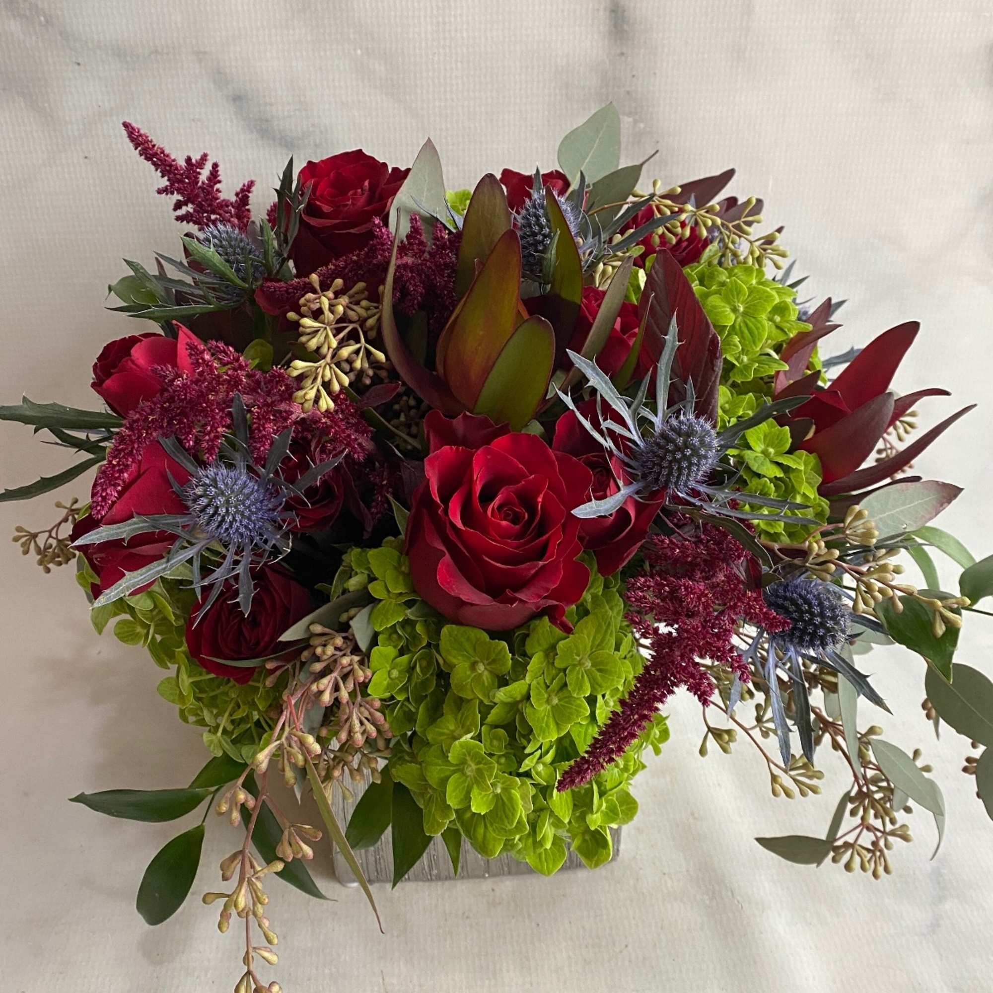 Red roses, green hydrangeas, blue thistle, seeded eucalyptus and hanging amaranths fill