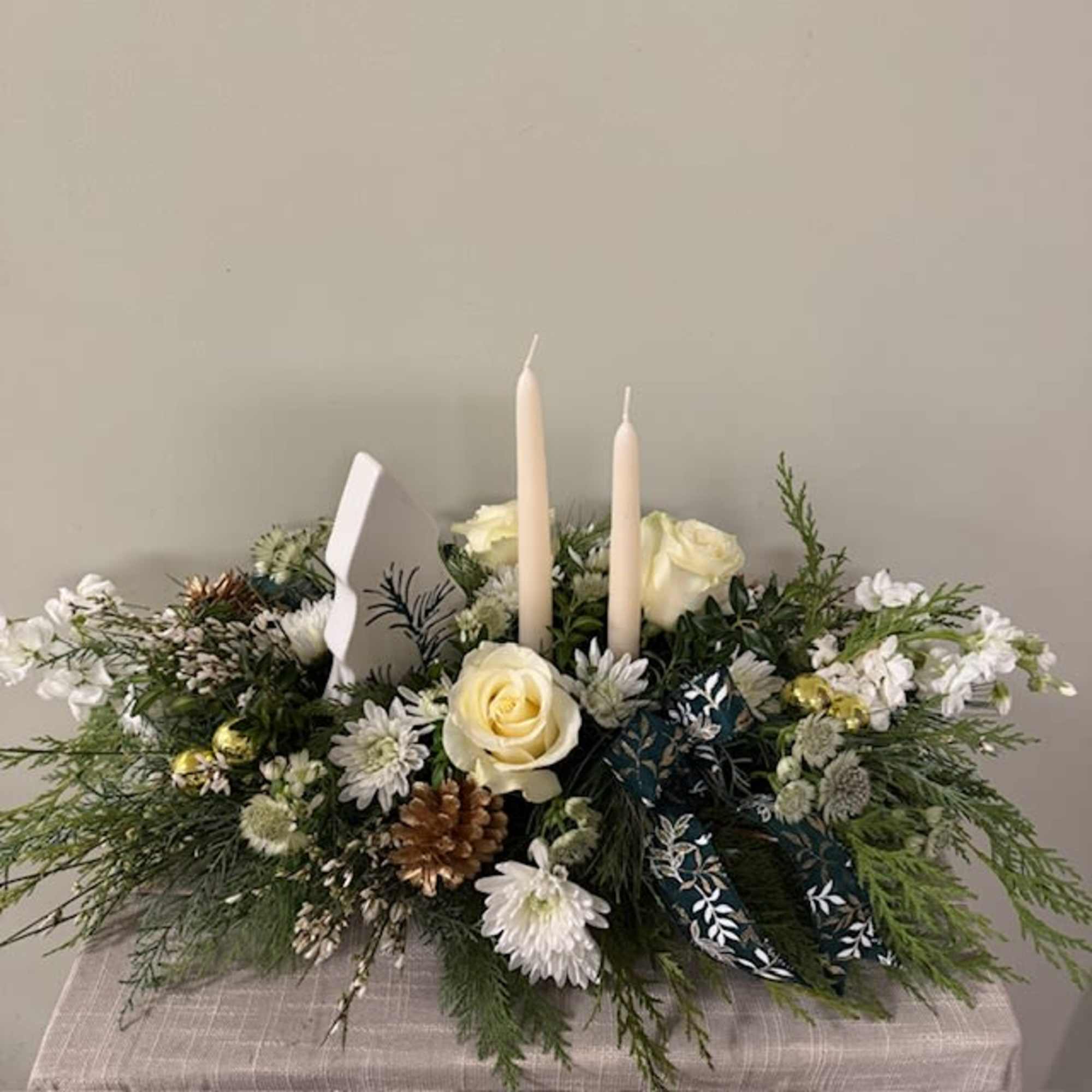 Double candle centerpiece includes winter greens, florals and a ceramic keepsake Christmas