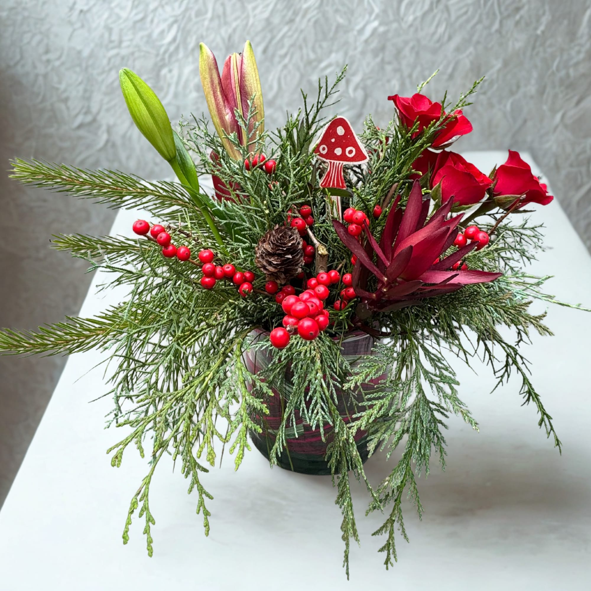 A cheerful holiday arrangement featuring rich evergreens, vibrant red roses, winterberries, and