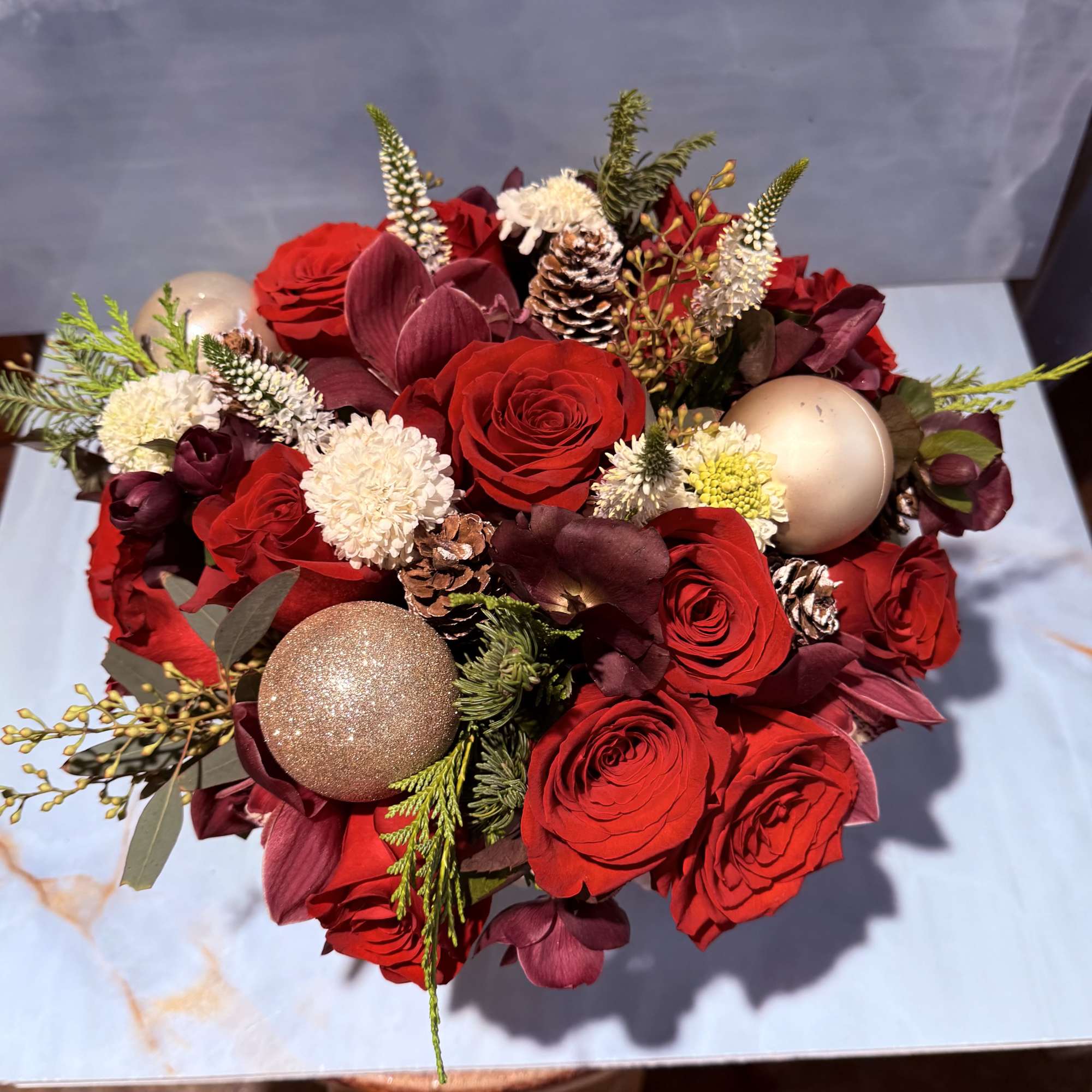 This arrangement consists of burgundy blooms, red roses, and evergreen foliage with