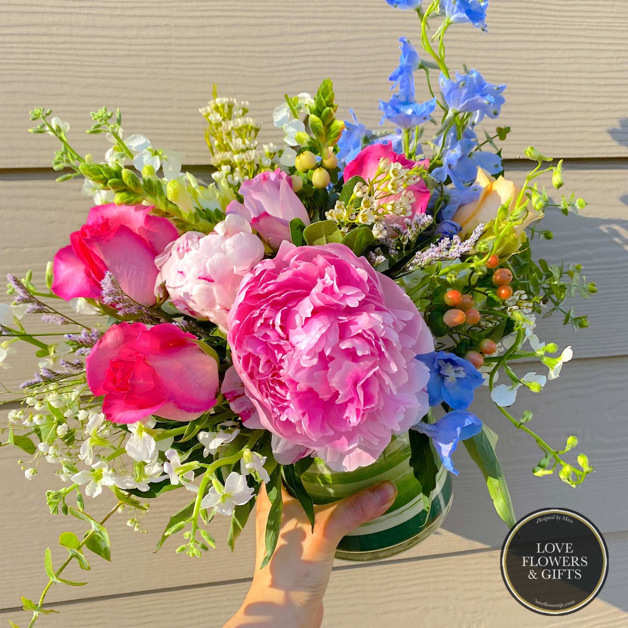 Bright bouquet of pink peonies, roses, and mixed blooms in a clear glass vase held in one hand