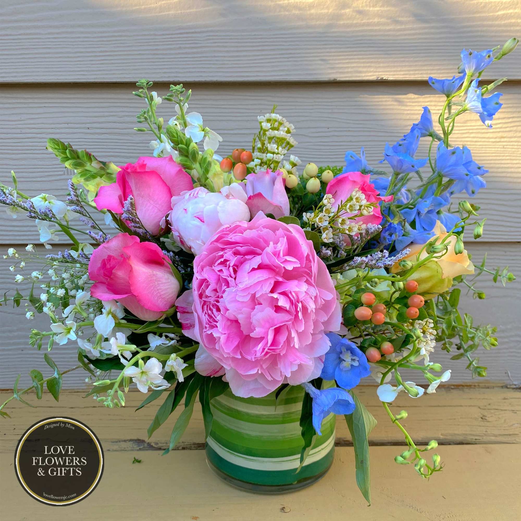 Short arrangement of pink peonies and roses with blue delphinium in a green striped vase