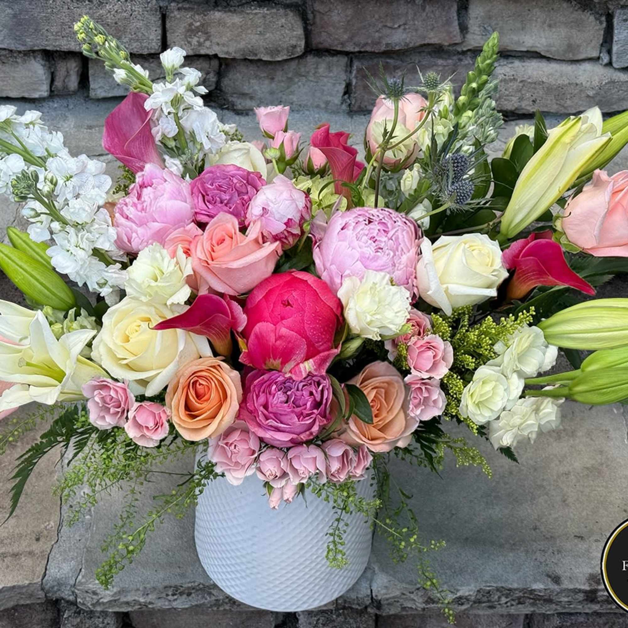 Lush arrangement of pink peonies, roses, lilies, and calla lilies in a white textured vase against a stone background