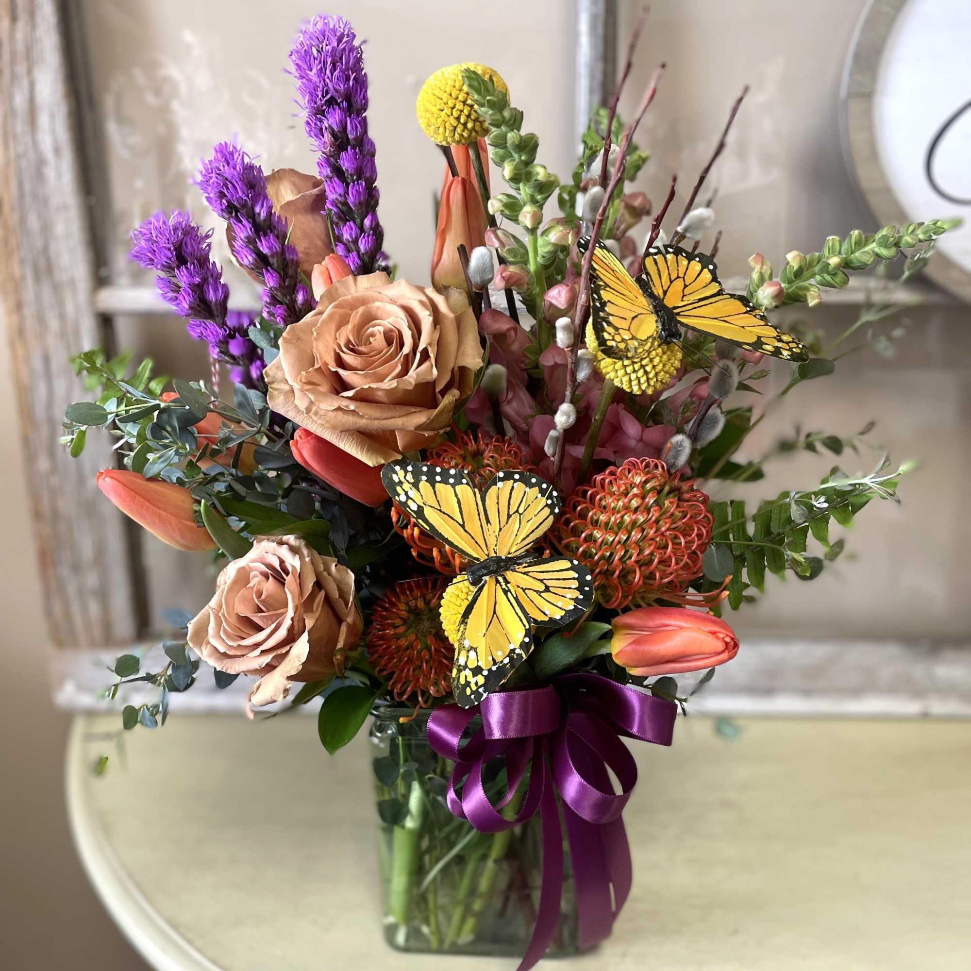 A lush, garden-style flower arrangement featuring roses, tulips, seasonal blooms, and decorative