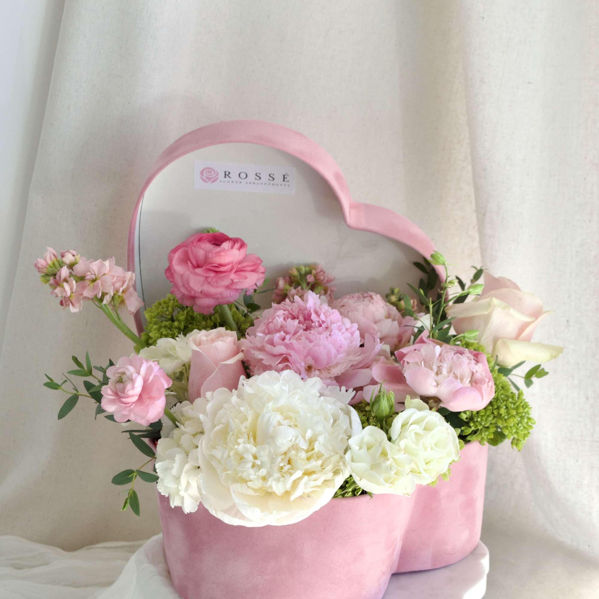 A heart-shaped pink velvet box filled with white, pink, and green flowers.