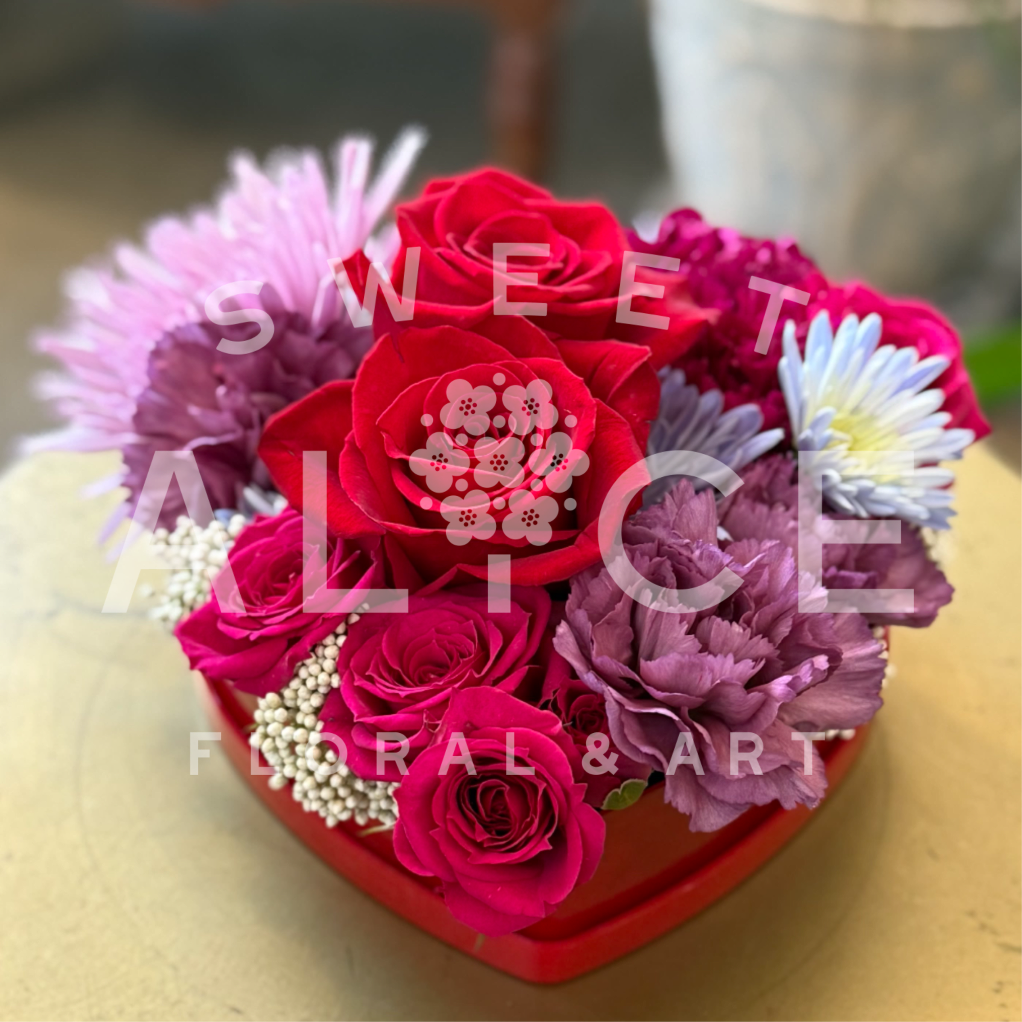 A red heart container, filled with red roses, sprays roses, carnations and