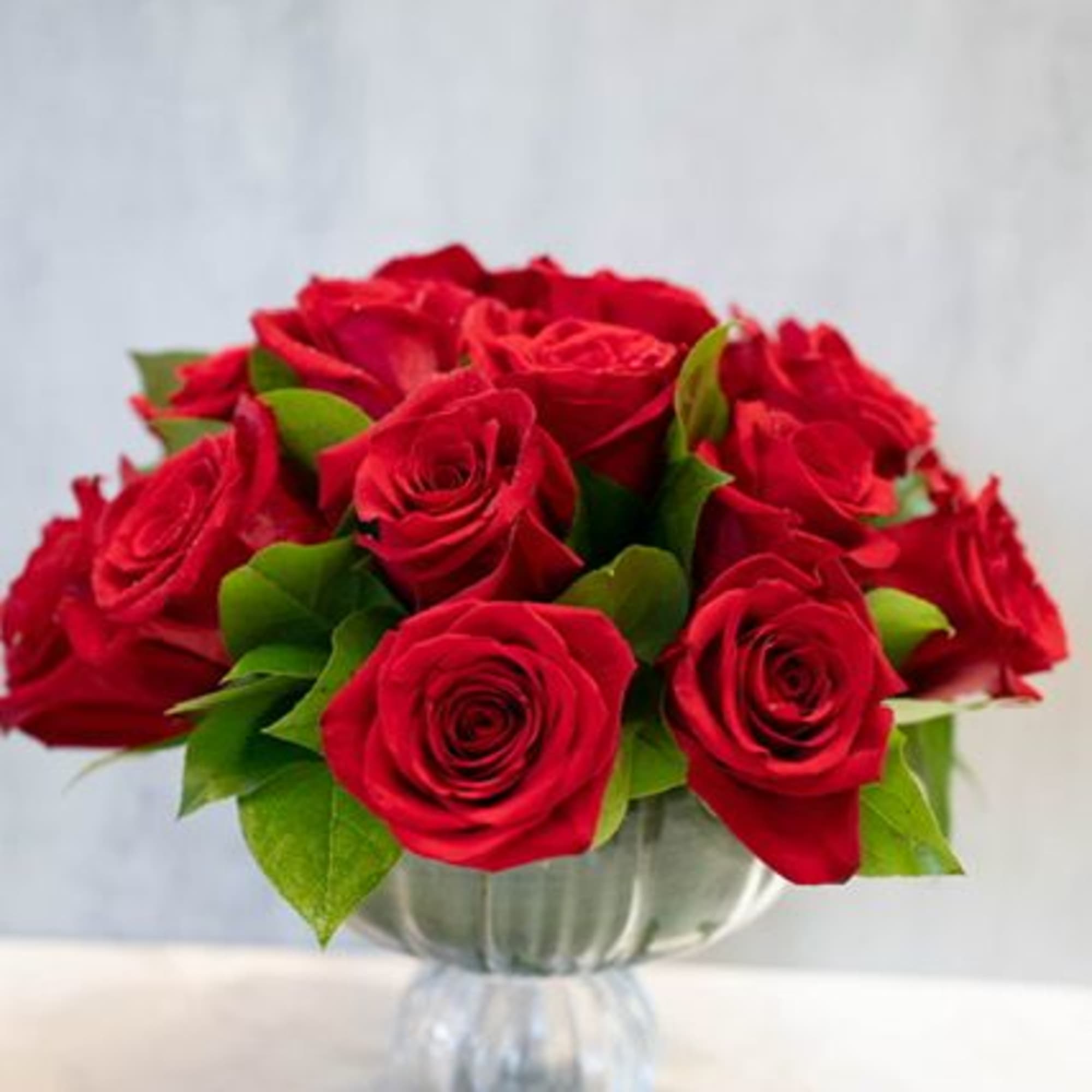 18 red roses designed with greenery in glass pedestal vase measuring 6.5"