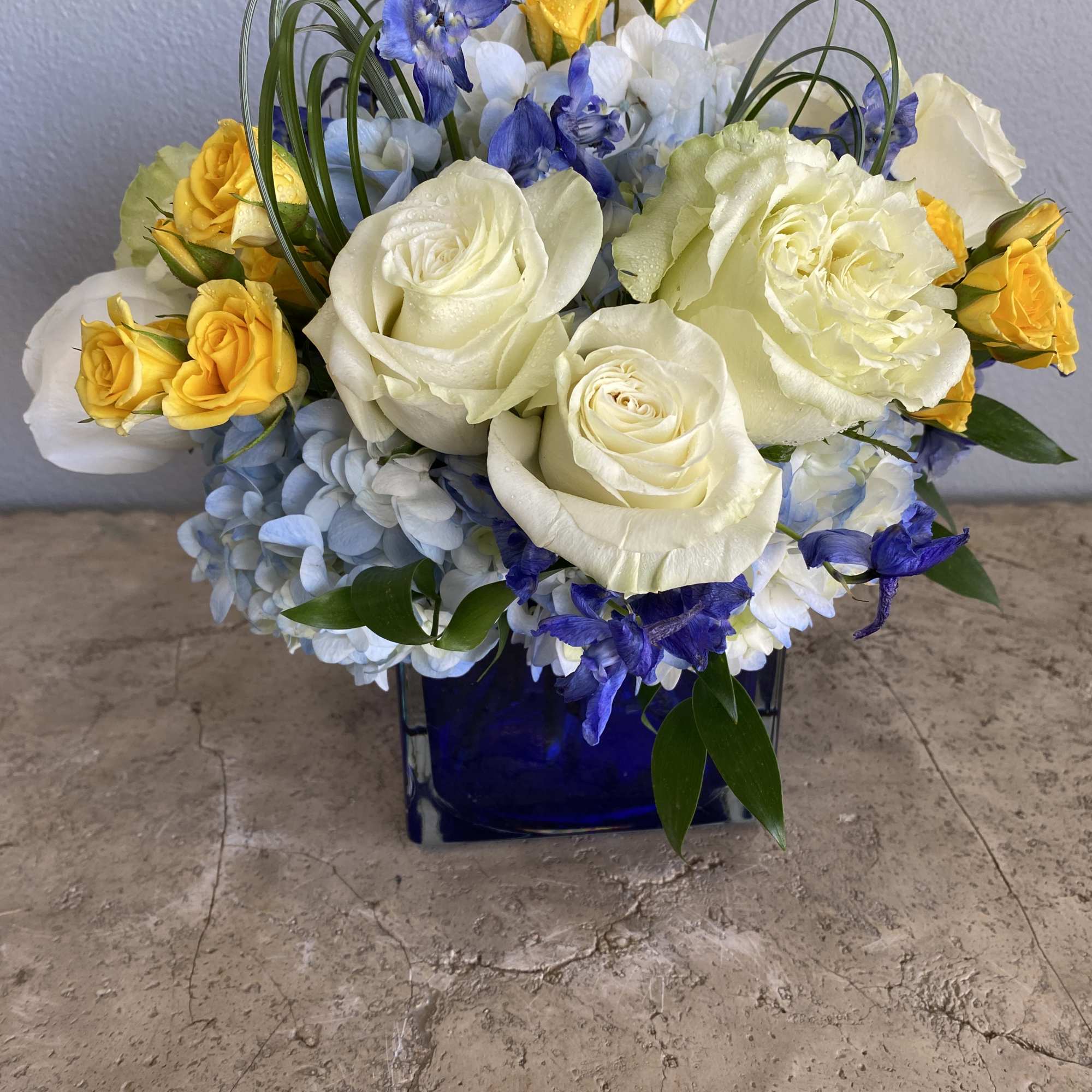 This bouquet features blue hydrangeas, blue delphinium, white roses, yellow spray roses