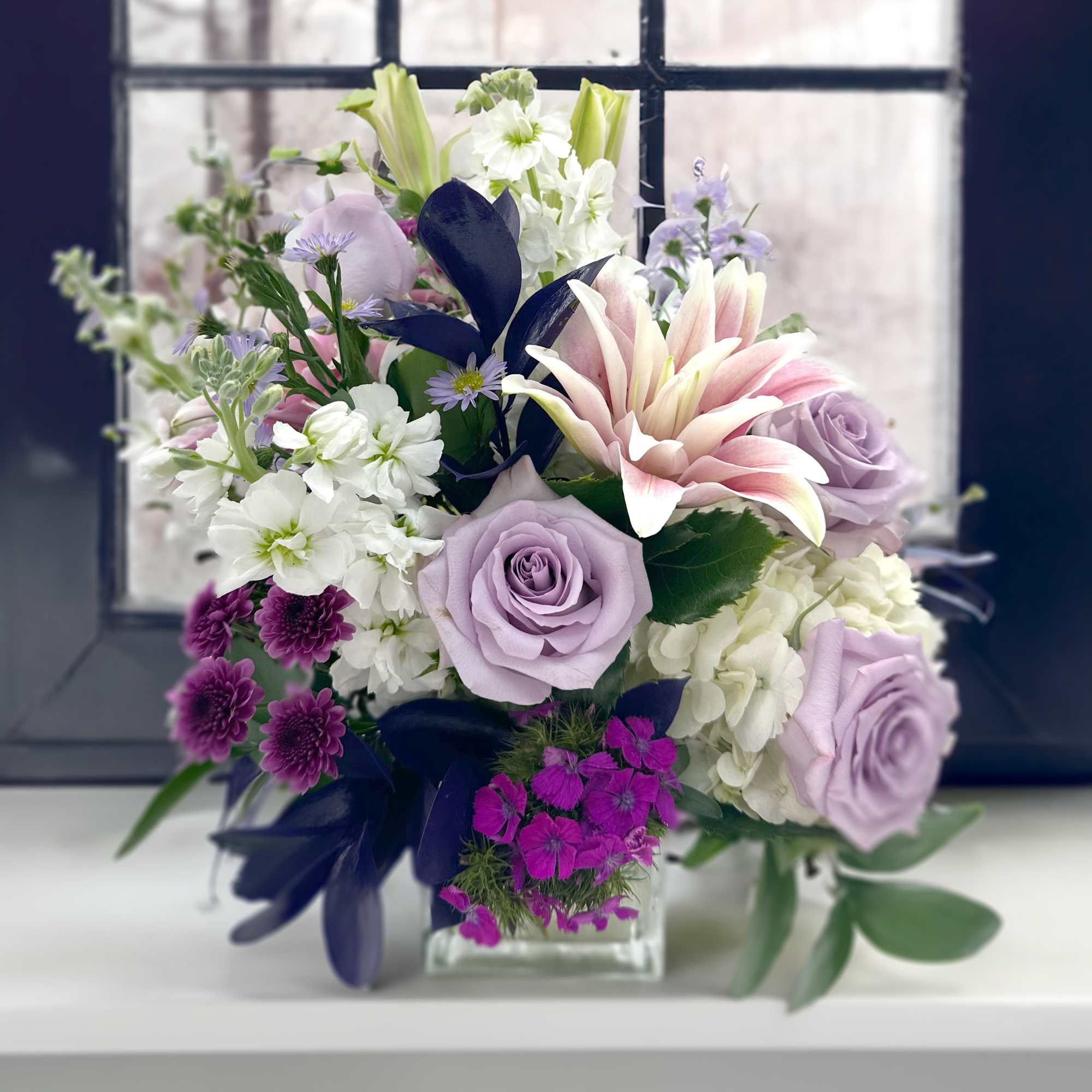 Enjoy getting lost in this arrangement of lavender roses, deep purple ruscus