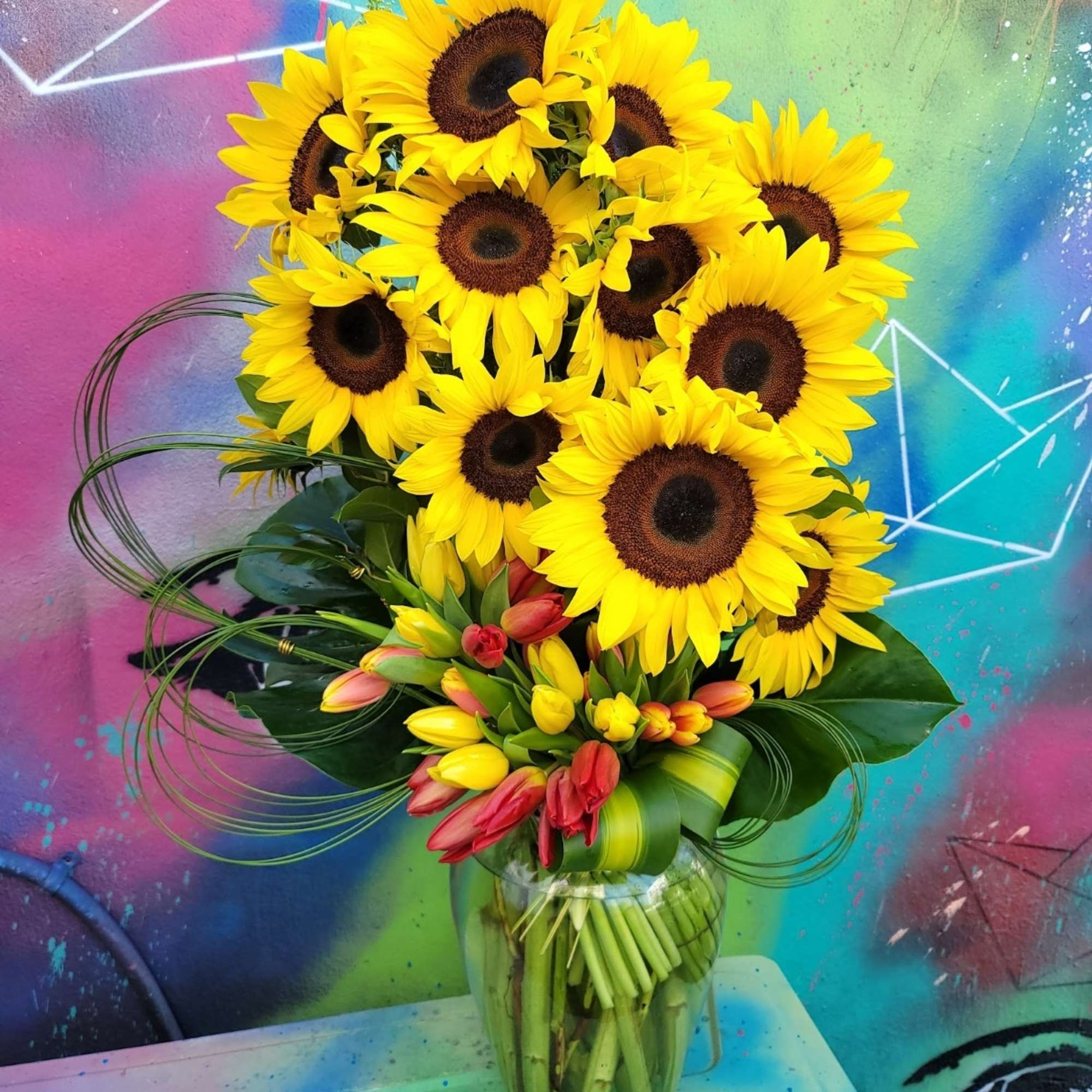 An arrangement in a clear vase made of sunflowers, Lillies, and greenery