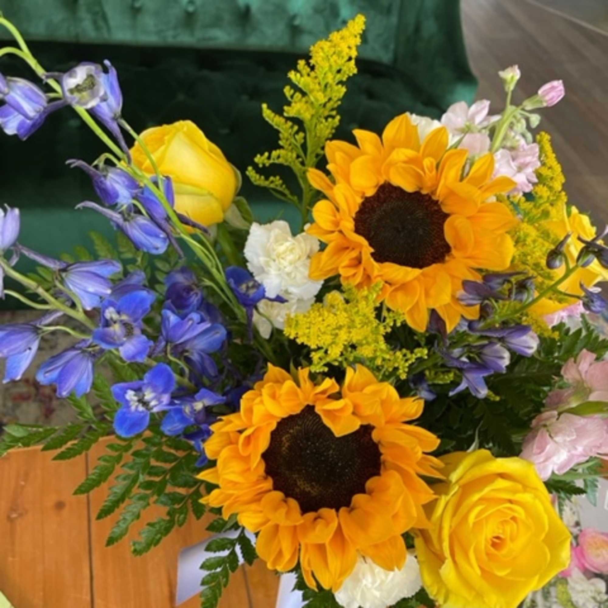 A country-inspired arrangement featuring sunflowers, yellow, roses, mini carnations, delphinium, stock, and