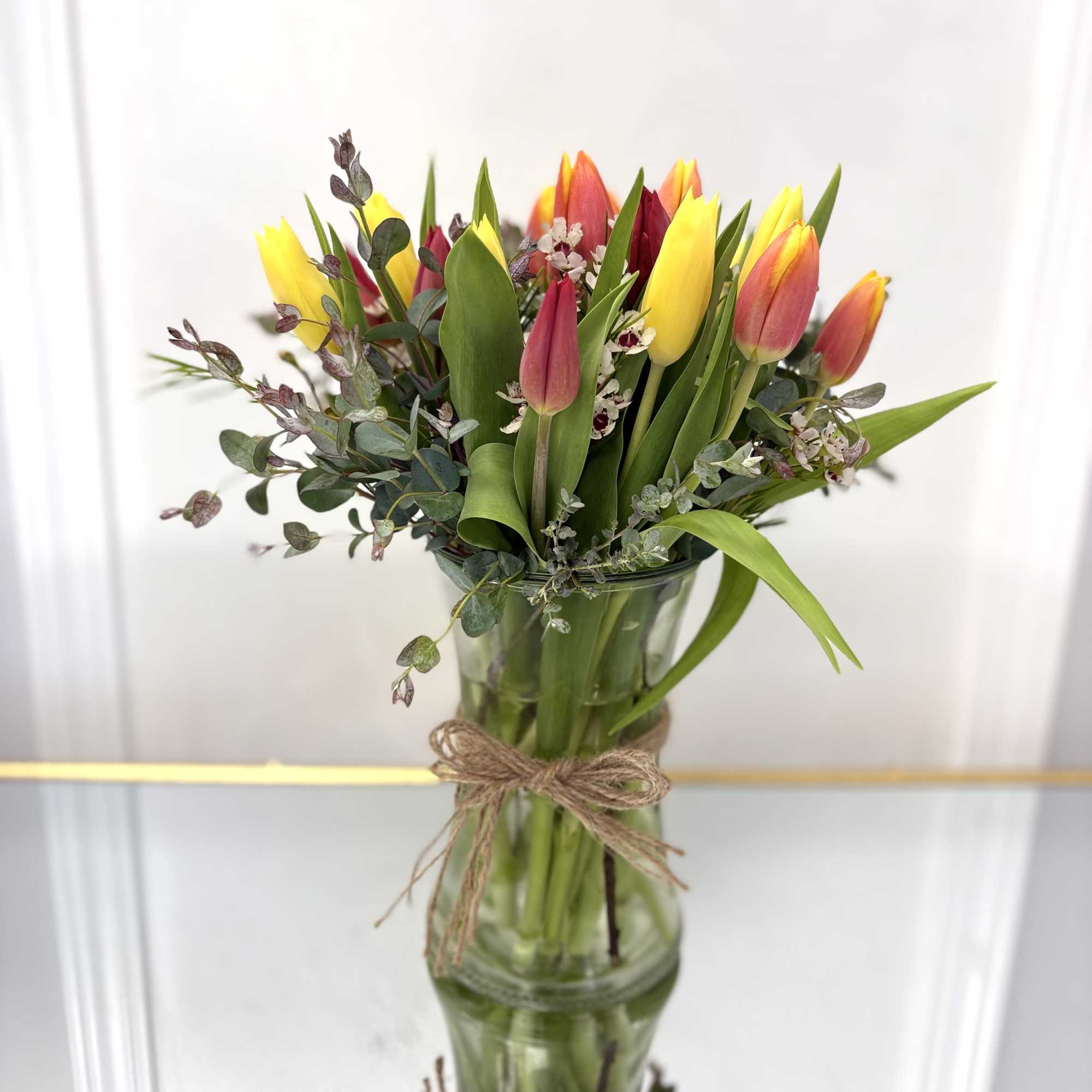 Yellow and red tulips with small white blooms in a tall clear glass vase tied with twine