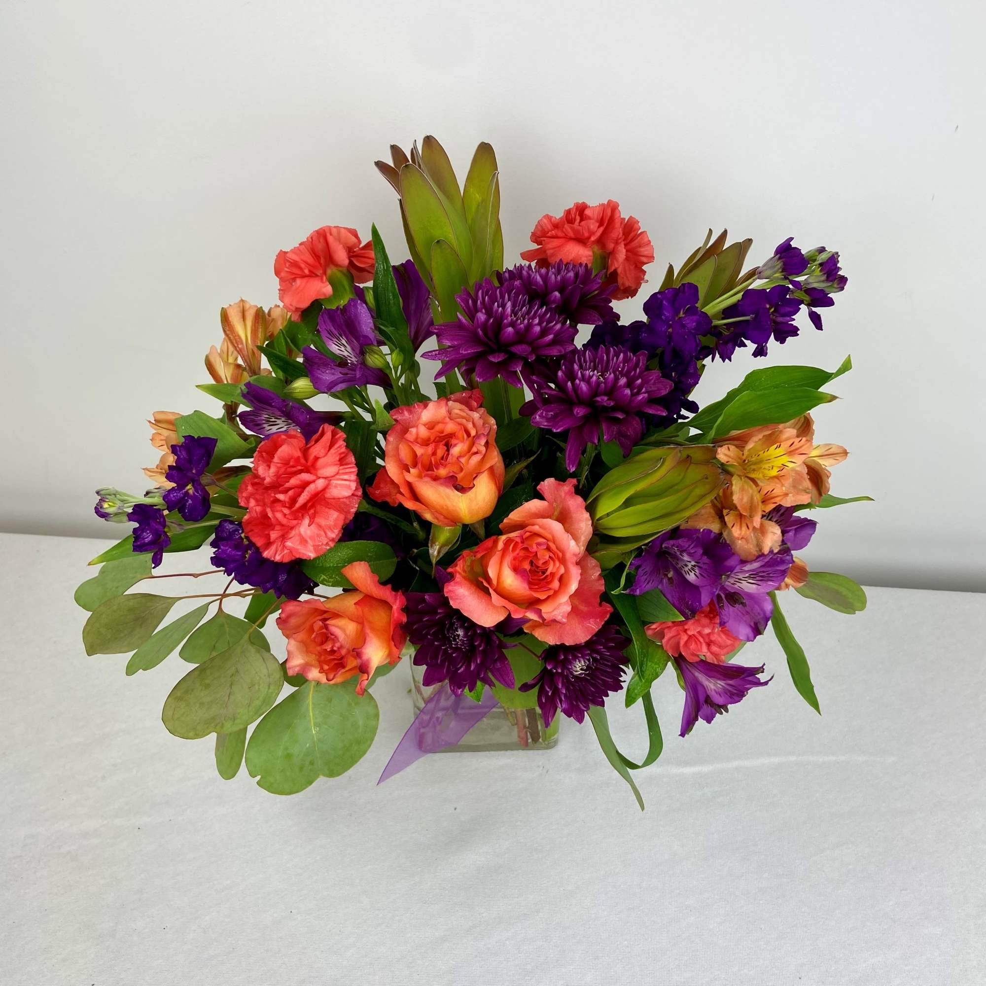 Mixed bouquet of orange roses, purple flowers, and coral carnations in a glass vase