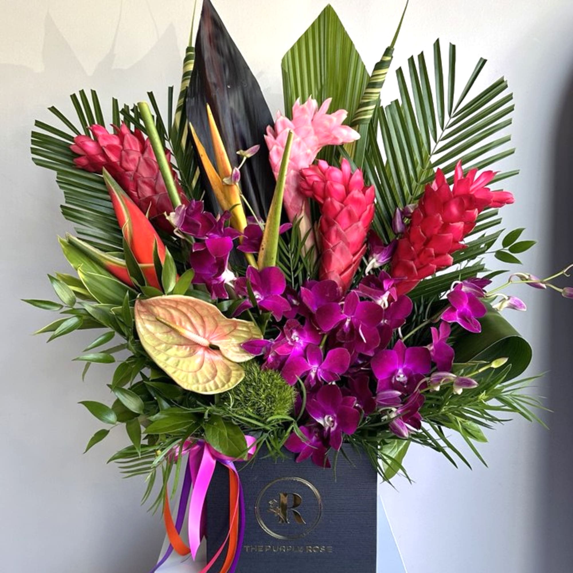 Tall tropical arrangement with pink ginger, purple orchids, and anthuriums in a black box with ribbons
