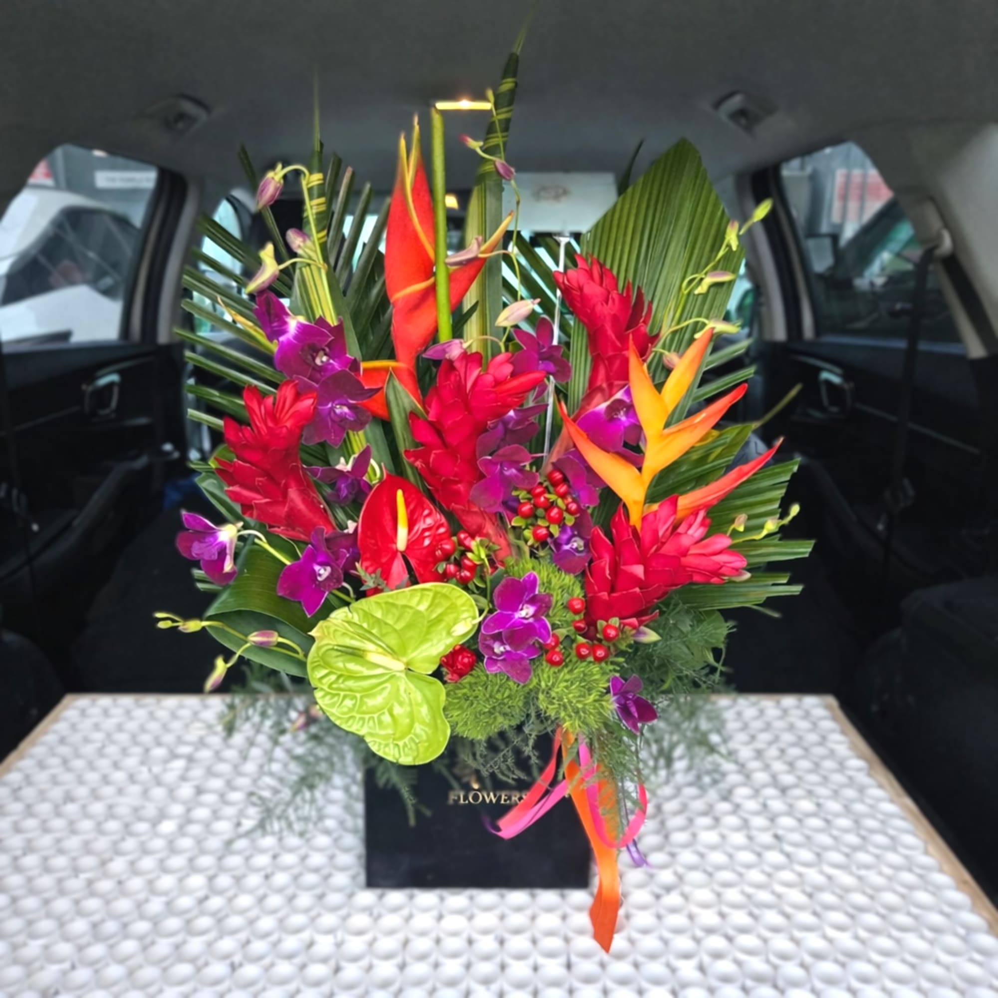 Tall tropical arrangement with red and orange blooms, purple orchids, and large palm leaves in a black box.
