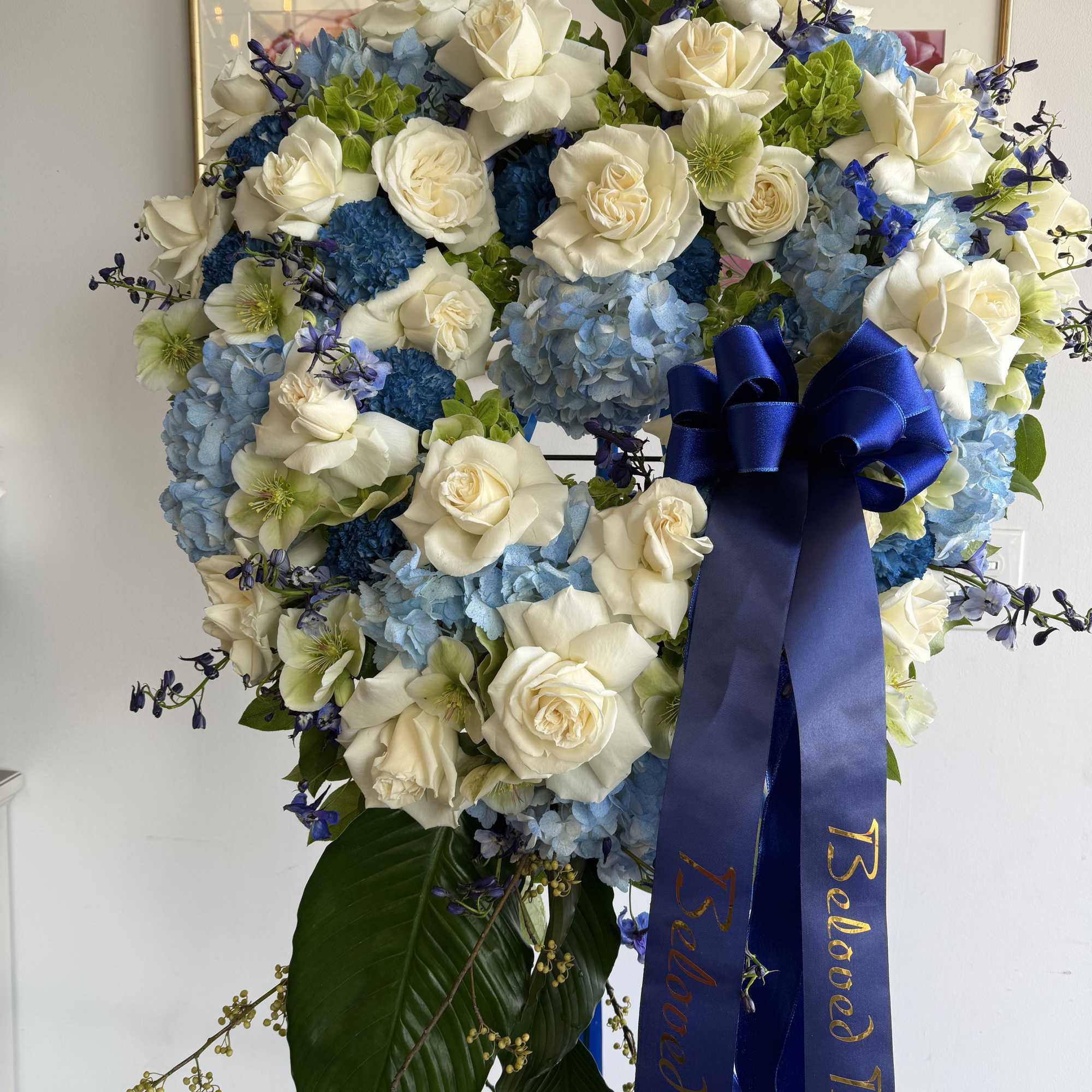 Standing wreath of white roses and blue hydrangeas with navy memorial ribbon on an easel.