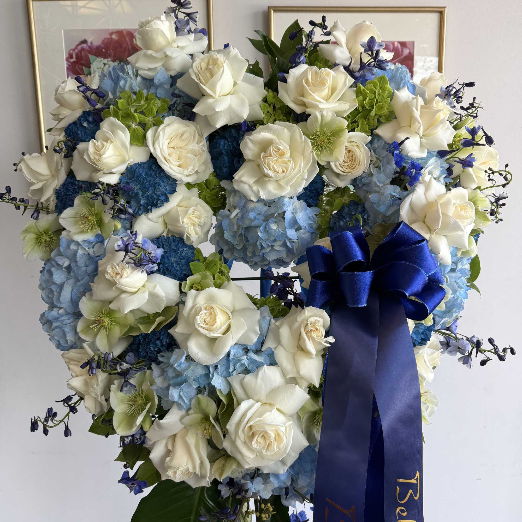 Heart-shaped wreath of white roses and blue flowers with a dark blue sympathy ribbon on a stand