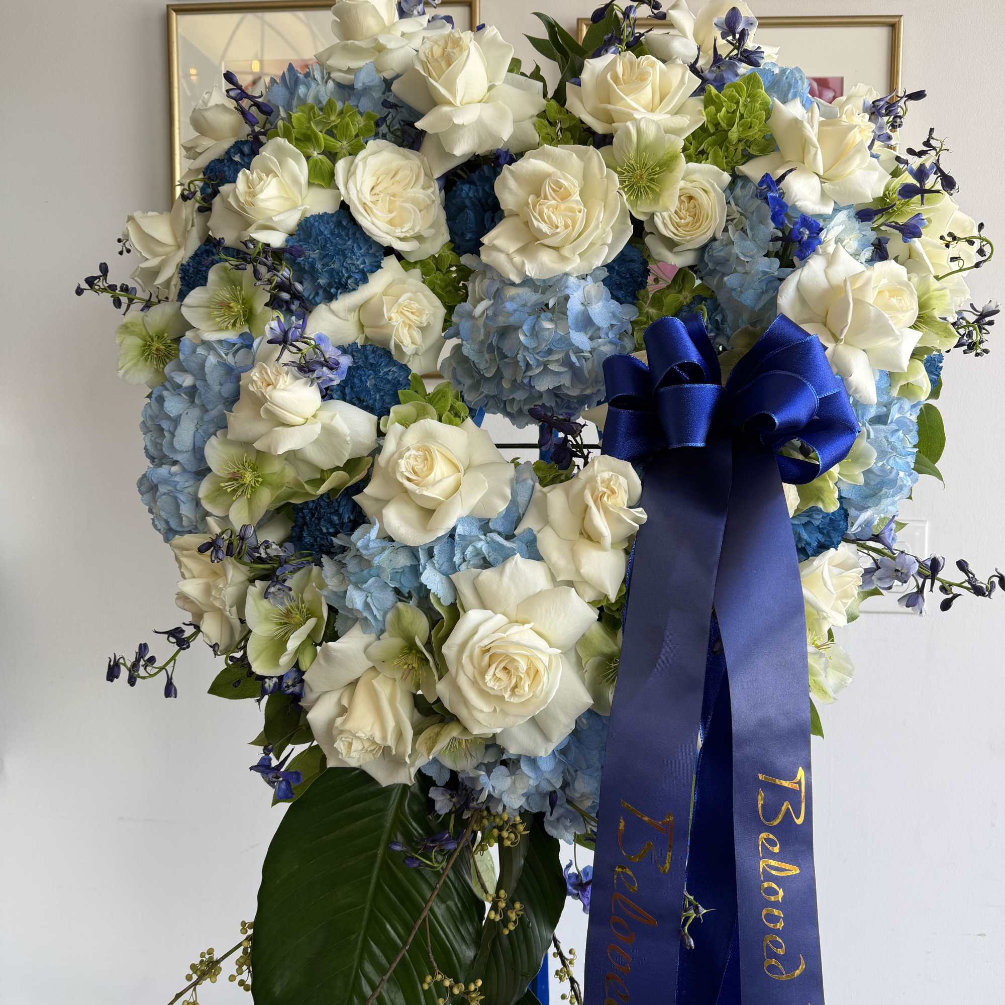 Heart-shaped standing wreath of white roses and blue hydrangeas with a blue sympathy ribbon.