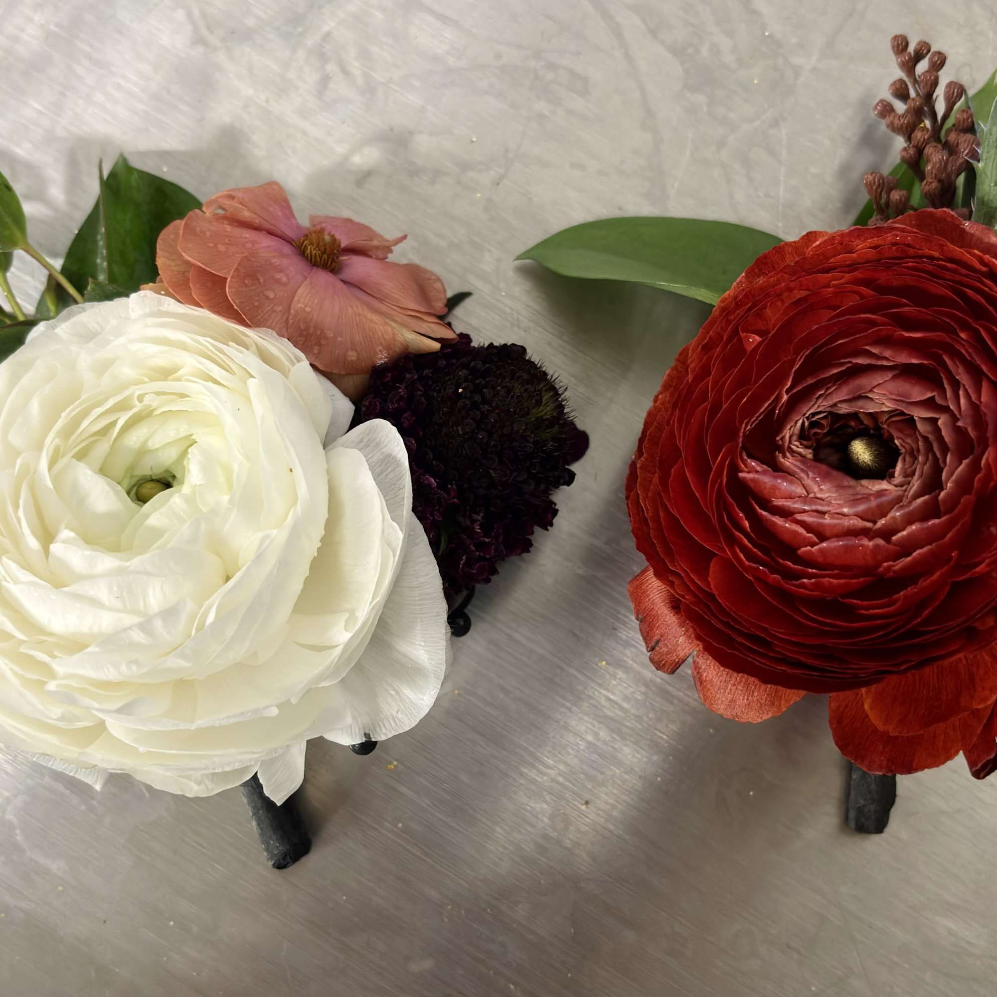 Two floral boutonnieres with white and red blooms on a metallic surface