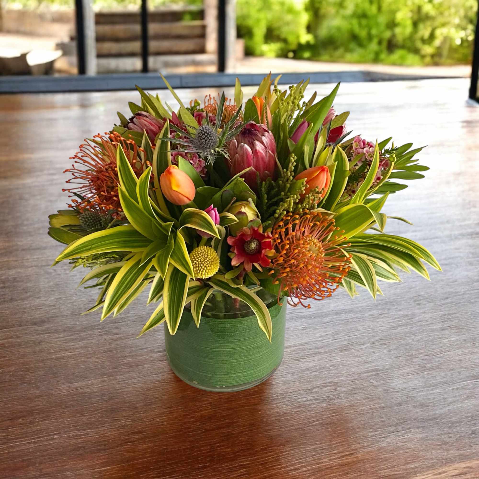 Low tropical arrangement with orange pincushion protea, tulips, and mixed blooms in a green-wrapped glass vase