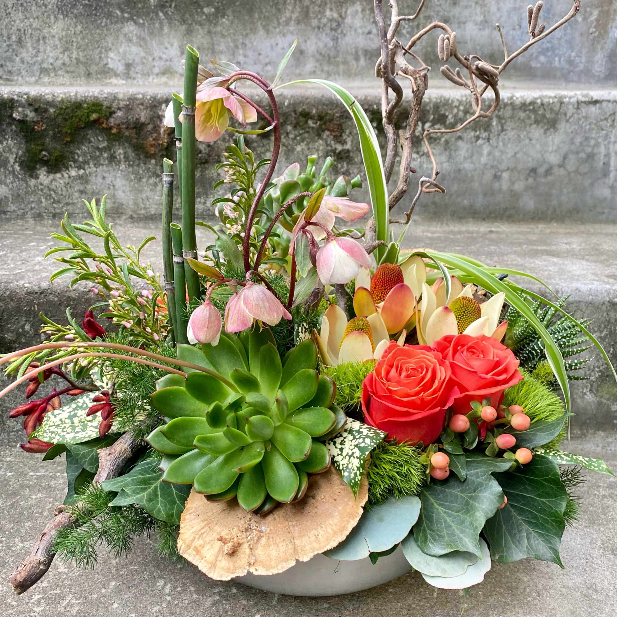 Succulent arrangement with pink hellebore-like blooms and red roses in a low bowl